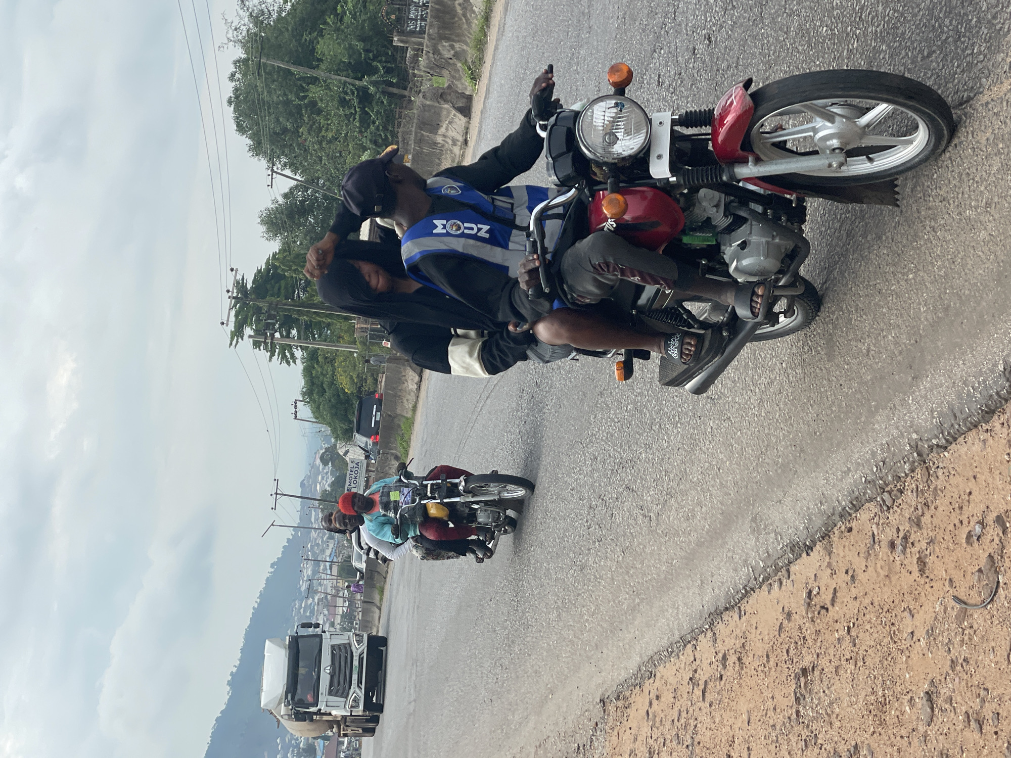 The image depicts a scene on a paved road in Lokoja, Nigeria, with a cloudy sky overhead. In the foreground, two people are riding a red motorcycle. The rider, wearing a dark baseball cap and a reflective blue vest with "MOUN" visible, is looking to the right. The passenger, with their face partially obscured by a black hooded garment, is seated behind the rider.
Further down the road, another motorcycle is visible, carrying three individuals. One person is wearing a turquoise top and a red beanie, while the other two are partially visible. To the left of the road, a large white truck with a cylindrical tank is traveling in the same direction. The roadside on the left is a dirt embankment with scattered stones. On the right side of the road, there is a concrete barrier and vegetation. In the background, buildings and hills are visible, suggesting an urban or semi-urban setting. A sign on the right side of the road indicates "MOTEL 5 LOKOJA".