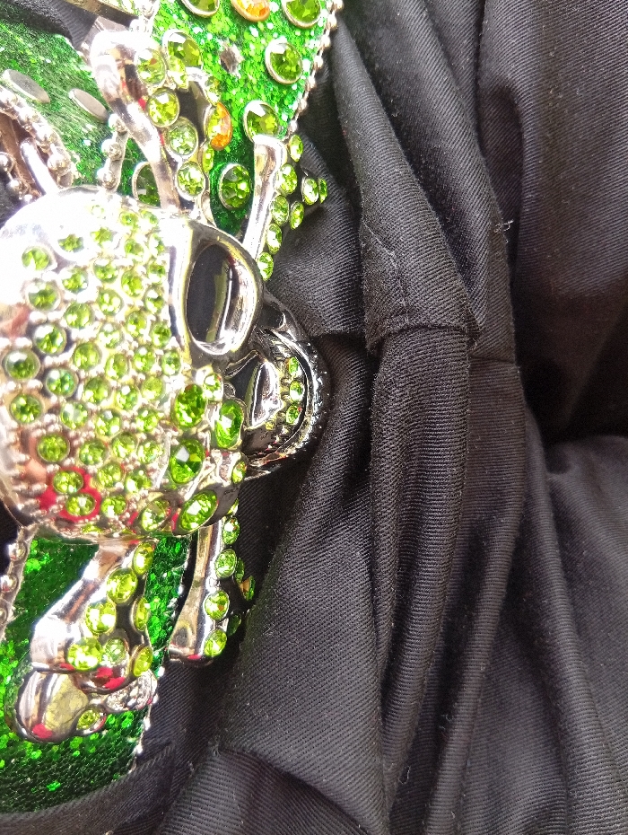 This close-up image features a decorative, silver-toned metallic object, likely a belt buckle or an emblem, set against a dark, possibly black, fabric background. The object is designed as a stylized skull and crossbones motif, heavily encrusted with numerous bright green rhinestones or gems that sparkle under the light. A few smaller orange gems are also visible among the green ones, particularly towards the upper right of the skull. The metallic parts of the skull, including the eye sockets and teeth, are highly reflective. The background fabric is textured and appears to be folded or crumpled, showing distinct diagonal lines from its weave. There are no people visible, and no specific activity is taking place; the image is a still life portrait of the object. No text is visible in the frame. The image was captured in Naro Moru, Kenya.