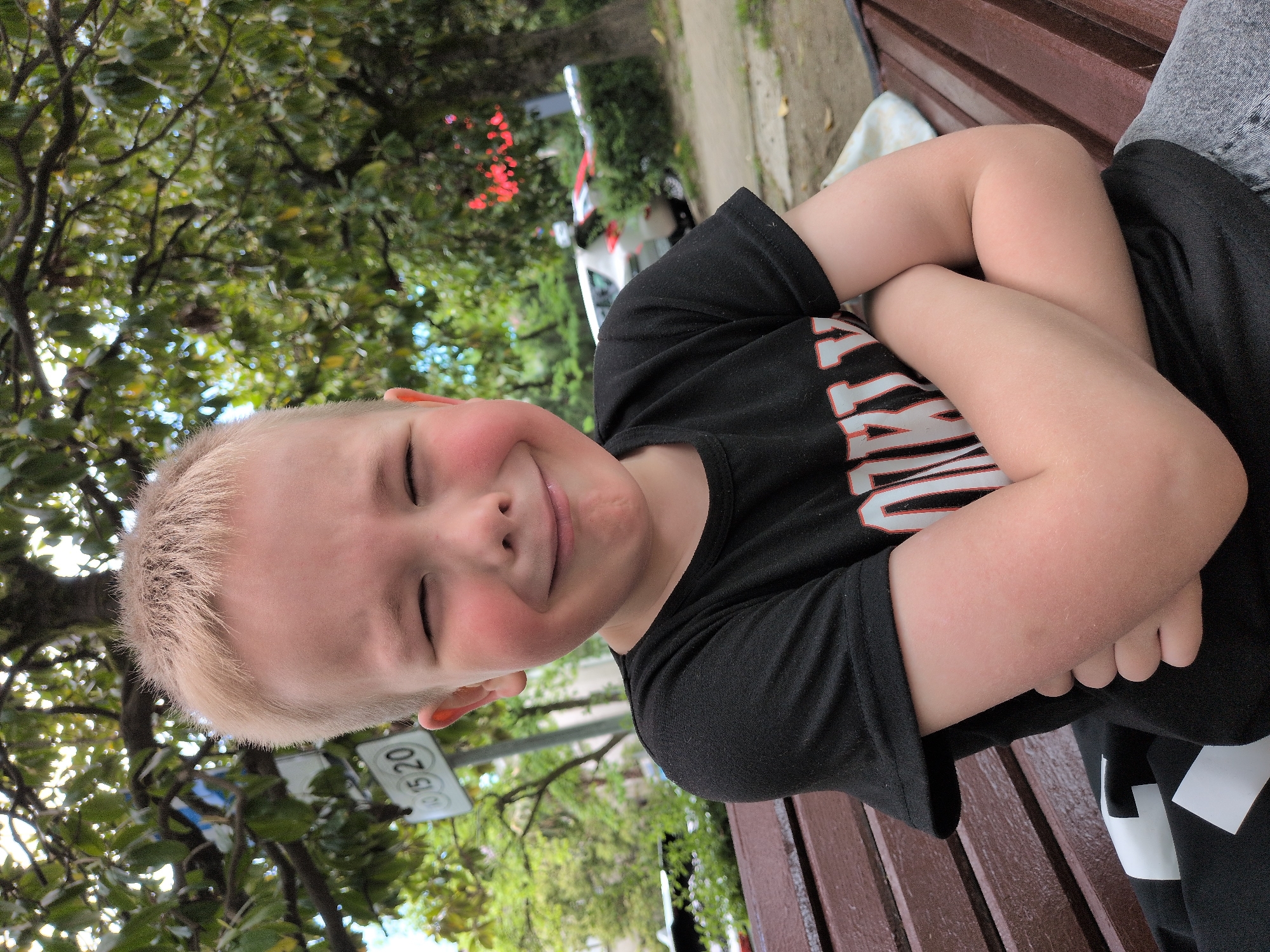 A fair-skinned, blond-haired boy with flushed cheeks is sitting on a brown wooden bench. He is wearing a black t-shirt with white lettering on the front. He has his eyes closed and is smiling slightly. His arms are crossed in front of him. The background includes trees with green foliage and a street sign that reads "10 15 20". There is a white vehicle with red accents visible in the background. The location appears to be in Sochi, Russia.