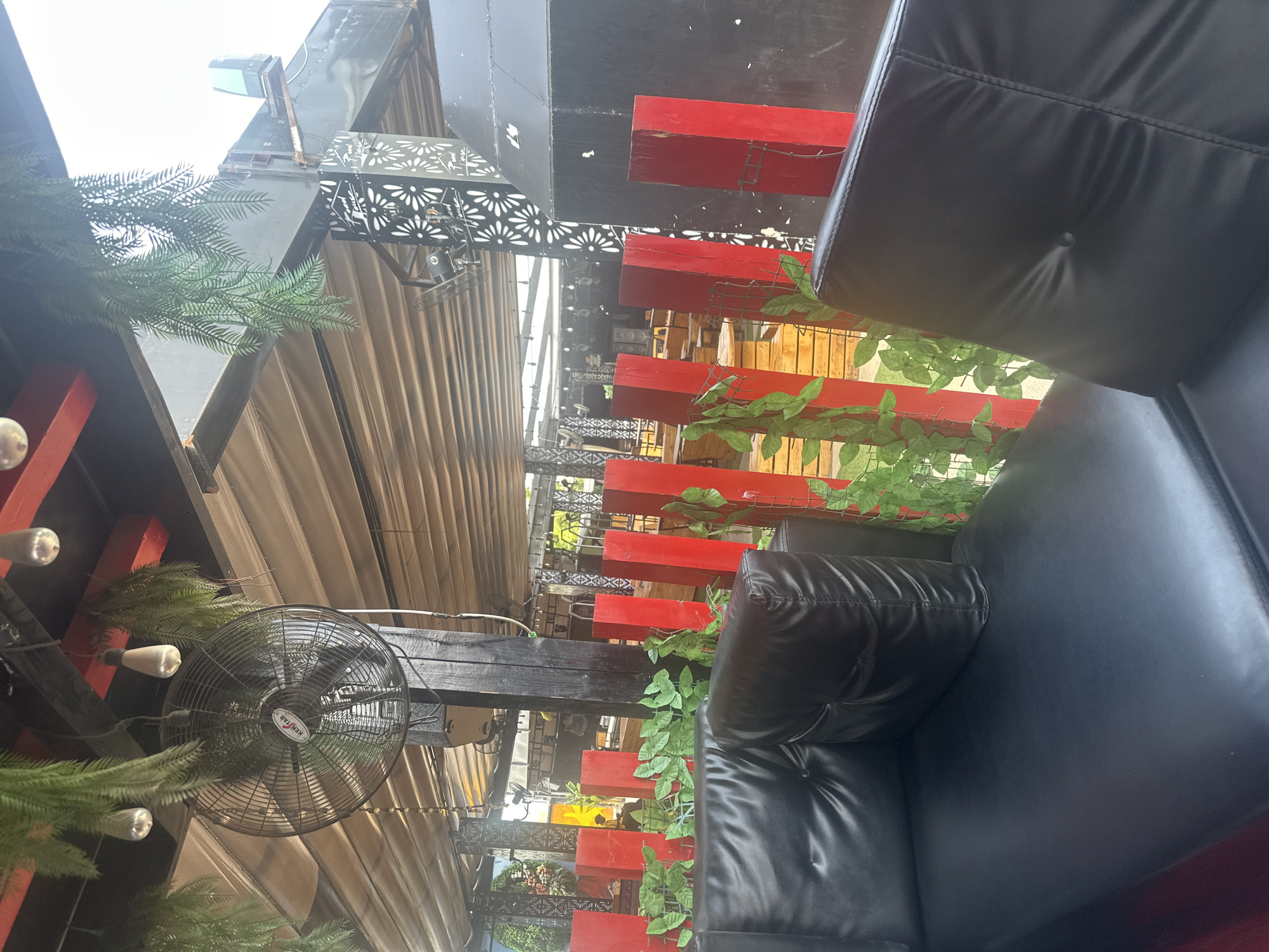 The image shows an outdoor seating area with black leather sofas. A black oscillating fan is mounted on a wooden beam above the seating. Artificial green foliage is draped around the seating area and along red wooden beams that form a decorative barrier. The setting appears to be a restaurant or cafe in Howayi, Nigeria. The lighting suggests it is daytime, possibly late afternoon given the soft light. The overall ambiance appears relaxed and comfortable. There is a visible brand name "KENSTAR" on the fan.