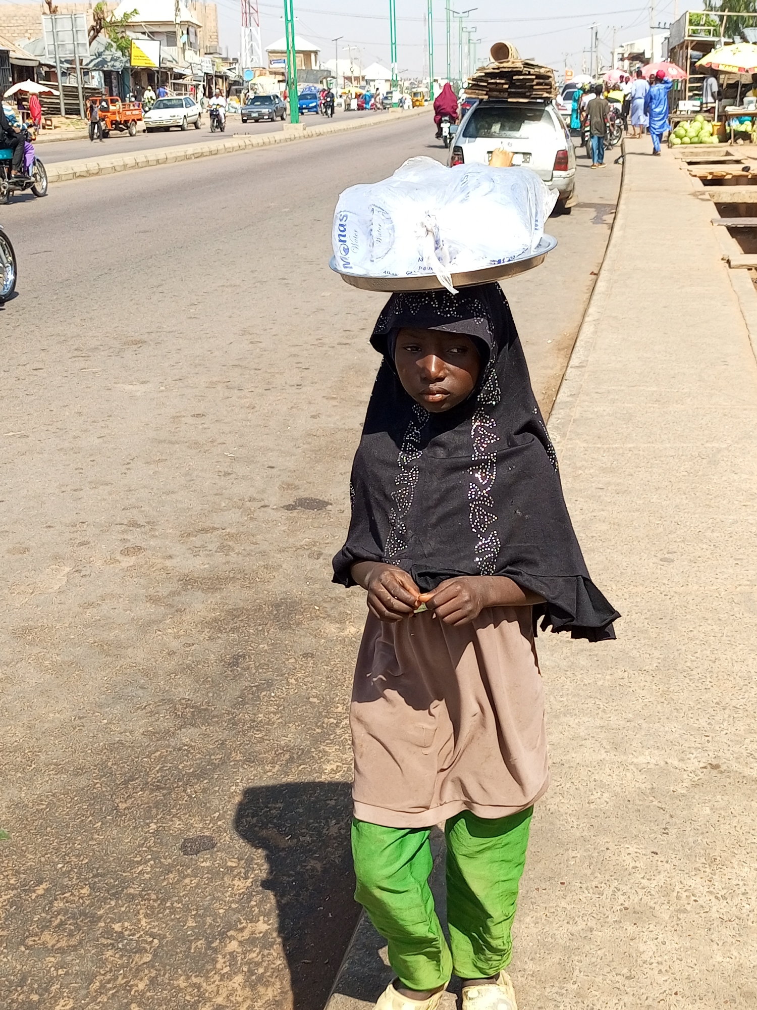 A young girl, likely under 10 years old, walks along a dusty road in Gombe, Nigeria. She is carrying a large metal tray on her head, laden with several white plastic-wrapped packages, possibly water bottles. The girl wears a black hijab adorned with shimmering rhinestones and a muted brown dress over bright green trousers. Her hands are clasped in front of her. In the background, a busy street scene unfolds with vehicles, including a white car overloaded with lumber, and various stalls lining the roadside. The overall impression is one of everyday life and commerce in a bustling urban environment.