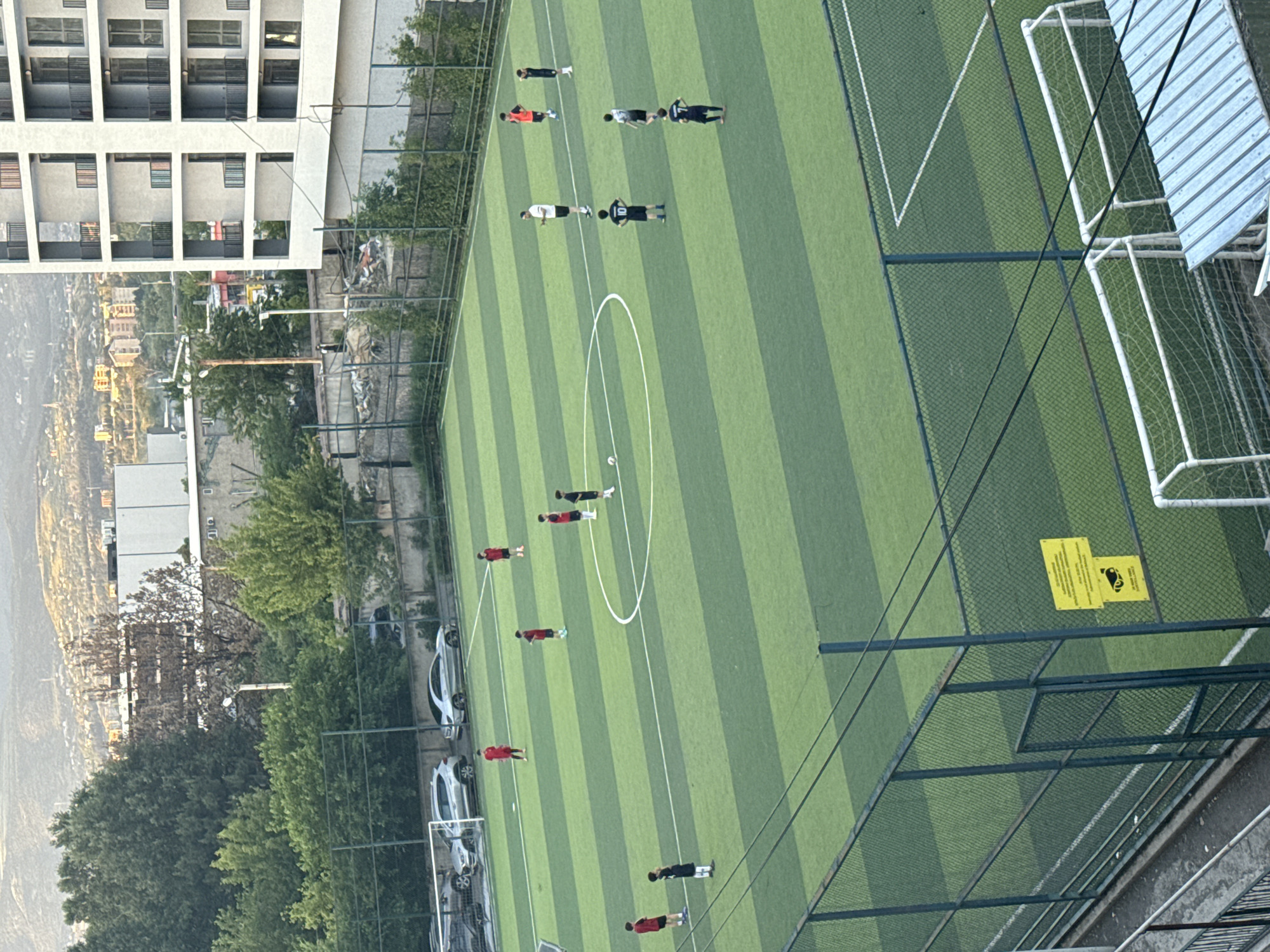 The image depicts an outdoor soccer field in Tbilisi, Georgia, with several young individuals, likely teenagers or pre-teens, engaged in a soccer training session. The field is artificial turf, marked with white lines, including a center circle and penalty areas. The players are scattered across the field, some in red jerseys and others in dark blue or black. One player is in the process of kicking a soccer ball. In the background, there is a multi-story apartment building, trees, and a distant cityscape with hills, indicative of Tbilisi's hilly terrain. A chain-link fence surrounds the field, and there are some parked cars visible beyond the fence on a paved area. The sky is overcast, suggesting it might be late afternoon or early evening. There is also a yellow warning sign attached to the fence.