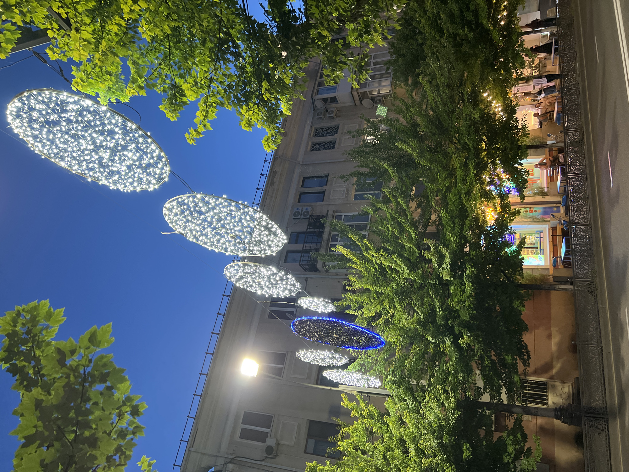 The image shows a street scene in Krasnodar, Russia, at dusk. Large, oval-shaped light installations made of white LEDs are strung across the street, illuminating the area. Trees with lush green foliage line the sidewalk, and behind them is a multi-story building with windows and air conditioning units. In the foreground, there is a street with cars and a sidewalk with a decorative metal fence. Several people are visible on the sidewalk, some sitting at tables outside what appears to be a cafe or restaurant. String lights are also visible over the outdoor seating area, creating a festive atmosphere. The sky is a deep blue, indicating the transition from day to night. The overall impression is of a lively urban evening setting.
