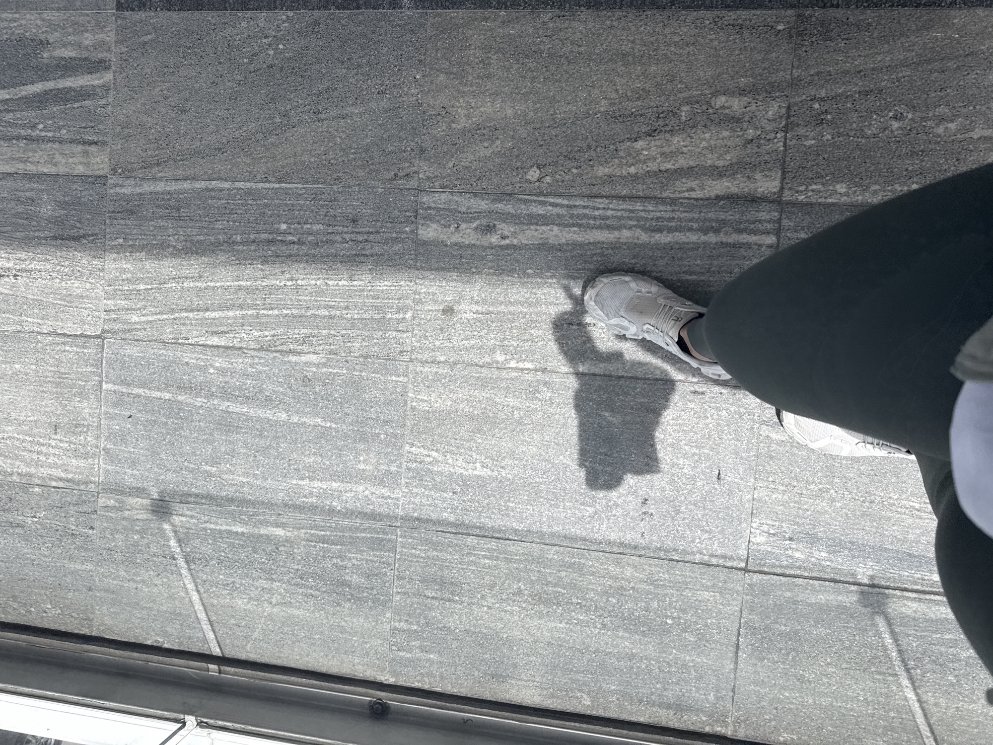 The image shows a view of a person standing on a gray granite tiled floor, likely a sidewalk, as indicated by the metal trim/edge visible at the bottom of the frame. The person is wearing gray sneakers and dark green leggings. The tiles have a linear grain pattern. There are no other people visible. The location is not identified in the image. There is no further information regarding location provided in the images.