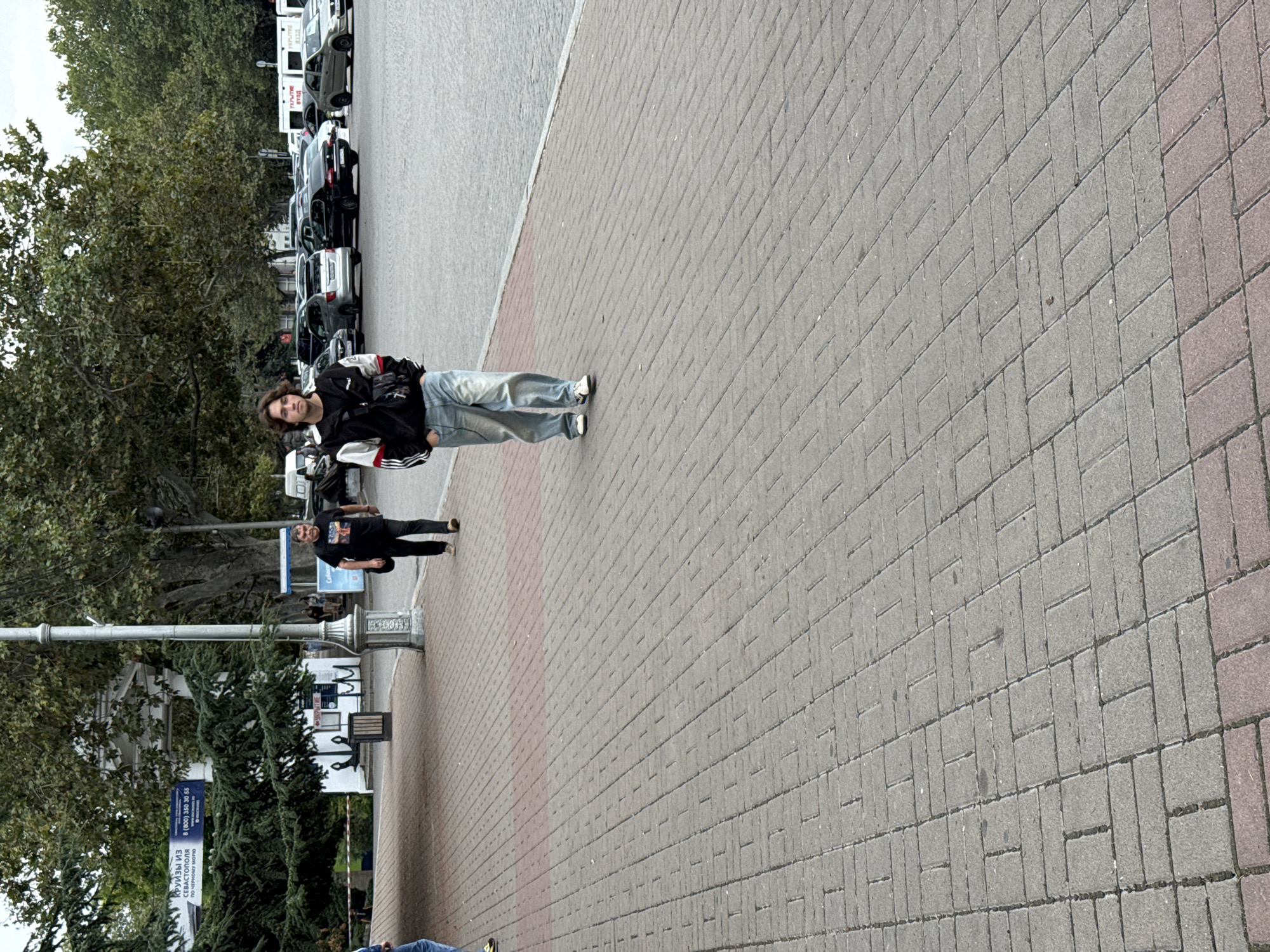 A young man walks on a paved sidewalk in Inkerman, Ukraine. He is wearing a black jacket with white and red stripes on the sleeves, light blue jeans, and white sneakers. He has a black crossbody bag slung over his shoulder and his hands are in his pockets. He is walking away from the camera and appears to be alone. In the background, there are trees, a parking lot with several cars, and a white building with a blue sign. Another man in a black t-shirt and dark pants walks further away on the same sidewalk.