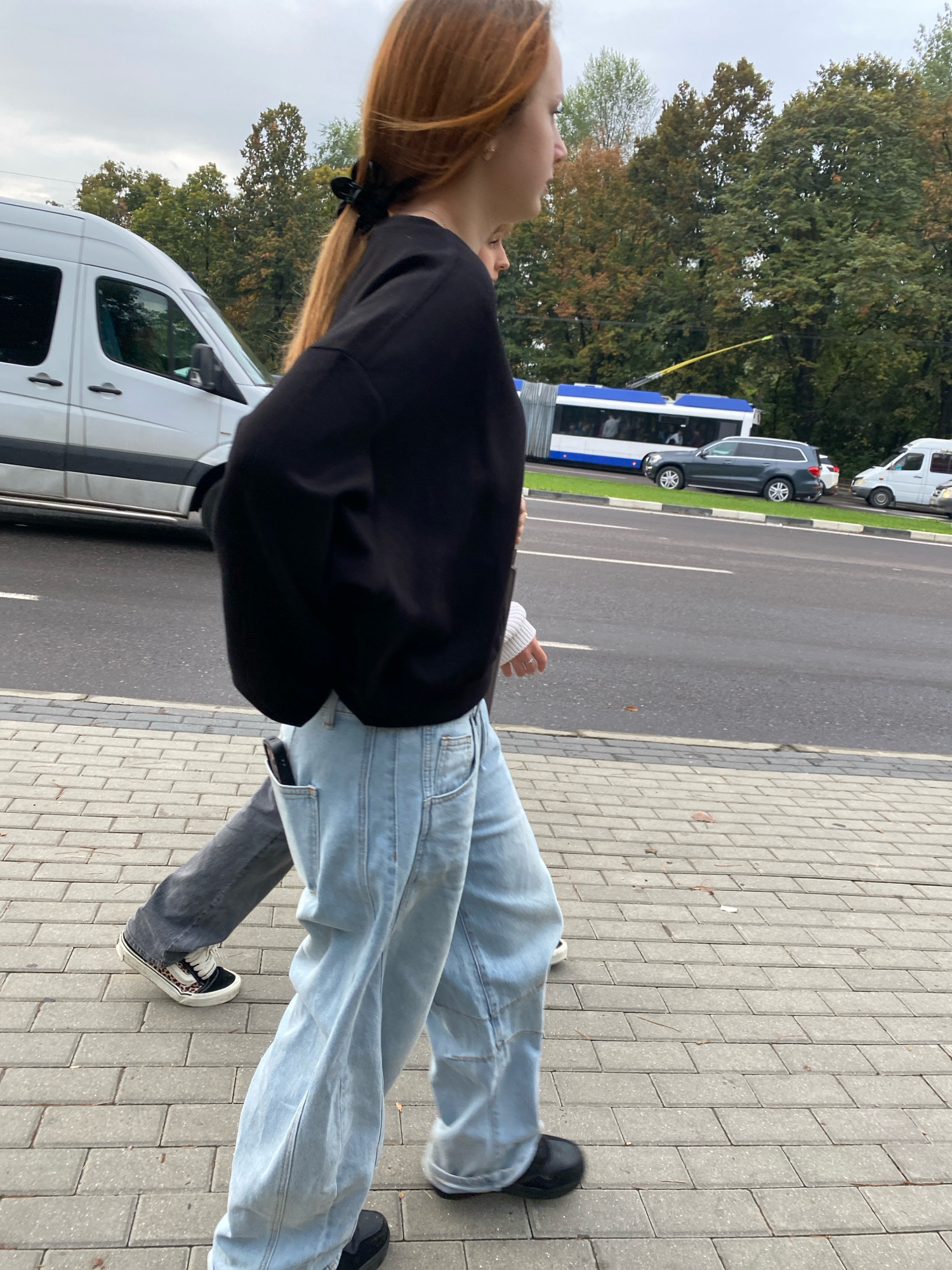 Two individuals are walking on a paved sidewalk adjacent to a street in Chisinau, Moldova. The person in the foreground, on the right, is a young female with long, reddish-brown hair tied back with a black scrunchie. She is wearing a black, oversized sweatshirt and light blue wide-leg jeans. A cell phone is visible in her right back pocket. She is also wearing black slip-on shoes. The second individual, to the left and slightly behind the first, appears to be wearing dark gray pants and black and white patterned sneakers. Both individuals are walking away from the camera.
The environment is urban, with a street visible to the right of the sidewalk, featuring a white van, a blue and white trolleybus, a dark gray SUV, and another white vehicle in the distance. Trees with green and some autumn-colored foliage line the street behind the vehicles. The sky is overcast. The sidewalk is constructed of light gray rectangular pavers laid in a repeating pattern.