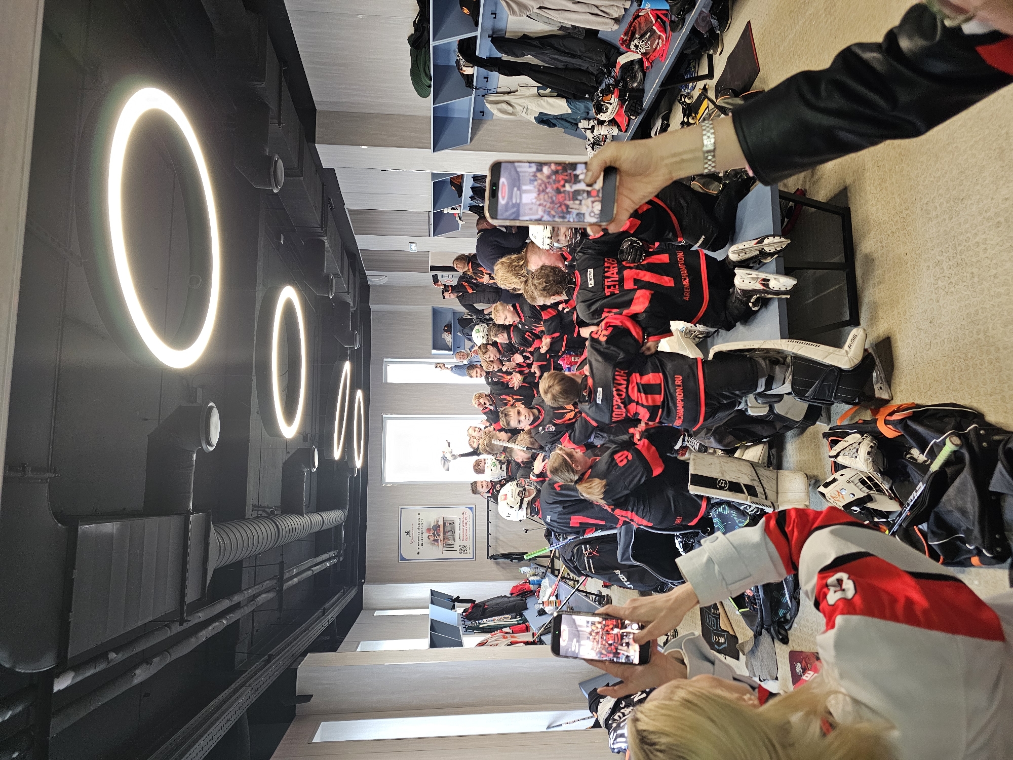 A group of young hockey players in black and red jerseys and hockey gear are seated on benches in what appears to be a locker room in the VTB Ice Palace in Moscow, Russia. The team is posing for a photo or video being taken by several people holding up smartphones. The locker room has blue shelves with hockey equipment and clothes stored on them. Overhead, there are distinctive circular light fixtures. Visible piping and ventilation systems are present near the ceiling. The locker room has a poster with Russian text on it.