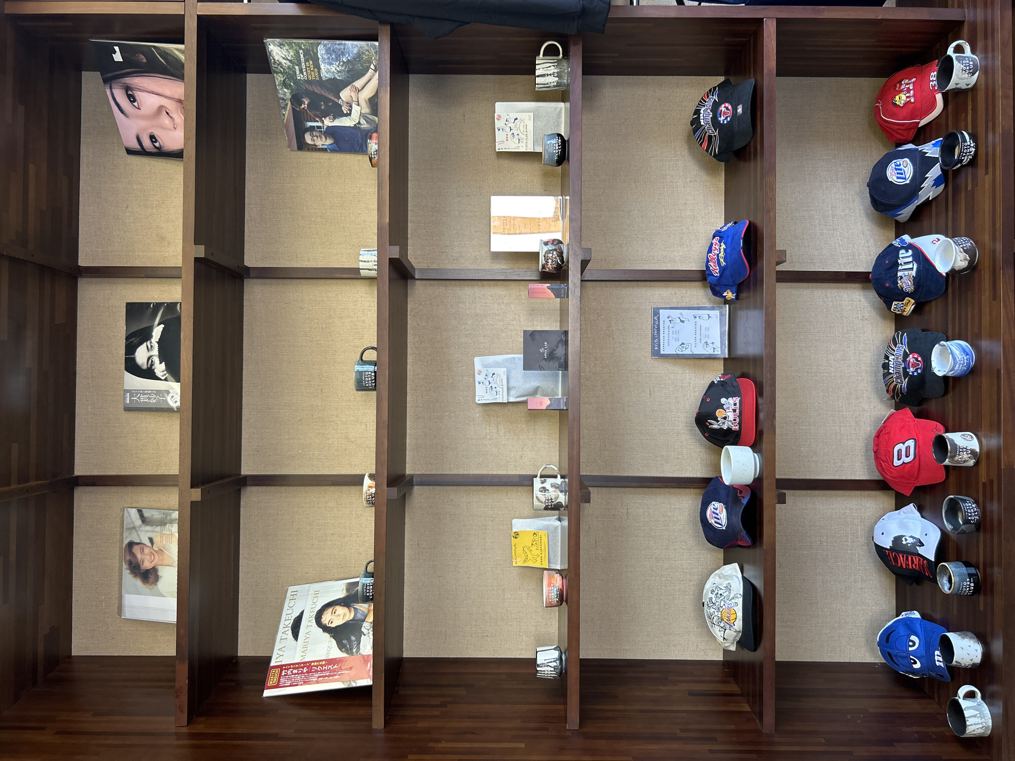 This is a photograph of a multi-tiered wooden shelf display containing various books, mugs, and hats. The shelves are lined with a textured beige fabric.
On the top shelf, from left to right, are three books. The first book is open, displaying a picture of a woman. The second book is square and appears to be a photobook of a woman. The third book is also a photobook featuring a woman's face.
The second shelf from the top holds two more books. The left book is a photobook with the title "MARIYA TAKEUCHI" visible. To the right of this book are two small mugs. Further to the right are two more books, one titled "KINGS OF CONVENIENCE" and another with a band name.
The third shelf from the top has a variety of items. On the left, there are two small decorative cups. Next to them is a small yellow bag with text on it. Following this are two mugs and two small packets or boxes. On the right side of this shelf, there are two small cups and a mug.
The fourth shelf from the top displays a collection of baseball caps. From left to right, there is a white cap with cartoon characters, a dark blue cap with "Miller" written on it, a red cap with "Bulls" and a cartoon character, and another cap visible on the far right edge. A white mug is placed between the first and second caps.
The bottom shelf also features baseball caps. From left to right, there is a blue cap with a cartoon character, a black and white cap with a portrait, a red cap with the number "8" and a cartoon character, and another cap partially visible on the right. In front of these caps, there are several small mugs or cups.
The overall environment suggests a retail display, possibly in a shop or gallery. The lighting is even and appears to be indoor ambient lighting. The background is out of focus, but the subject matter is sharp.