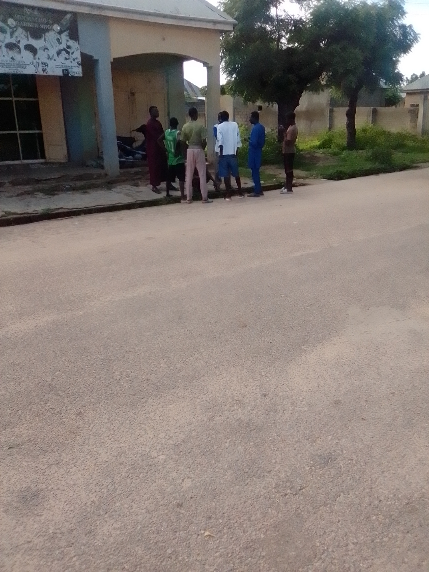 The image shows a group of six individuals standing on a paved road in front of a building. The building appears to be a shop, as indicated by a sign on its facade that reads "MICHAKO'S BARBER SHOP." Three of the individuals are wearing t-shirts and shorts, while the others are wearing traditional clothing. The environment is urban, with a road, a building, and some vegetation visible. The overall impression is a casual street scene in a Nigerian city, likely Gombe, given the presence of a barber shop and the style of clothing worn by the individuals.