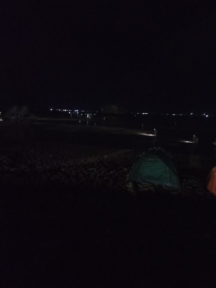 This image captures a dark, nighttime scene, likely a campsite, in Kecamatan Pujut, Indonesia. The primary subject in the foreground is a green camping tent, pitched on what appears to be sandy or rough terrain. To the right of the green tent, a portion of another tent, brownish-orange in color, is visible.
The scene is dominated by darkness, with very limited light sources. In the mid-ground and background, several faint, scattered points of artificial light suggest the presence of other individuals or activities. Some of these lights appear to be held by people, possibly using flashlights as they move around the dark landscape. Further in the distance, a line of brighter, more continuous lights can be seen, which could indicate a distant town, village, or a more populated section of the camping area.
The overall activity depicted is camping or an outdoor gathering at night. The time of day is clearly after sunset, with a completely dark sky and no visible natural light sources like the moon or stars. The weather conditions cannot be determined, but the atmosphere appears calm. There is no visible text in the image. The mood conveyed is one of tranquility and isolation, characteristic of a night spent outdoors away from significant light pollution.