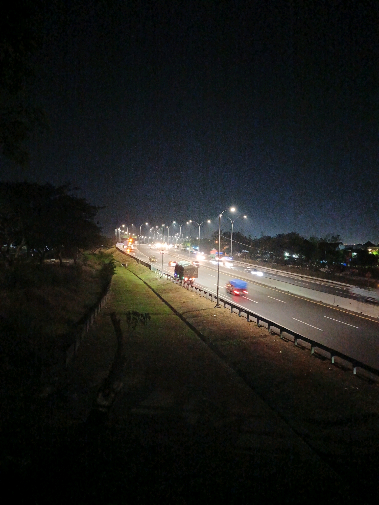 A nighttime aerial view shows a highway in Kludan, Indonesia. The highway is illuminated by streetlights, and several vehicles are visible on the road, including trucks and cars. The highway has multiple lanes and is bordered by a grassy median and a concrete barrier. Trees and vegetation are visible on either side of the highway, and buildings can be seen in the distance. The sky is dark and clear.