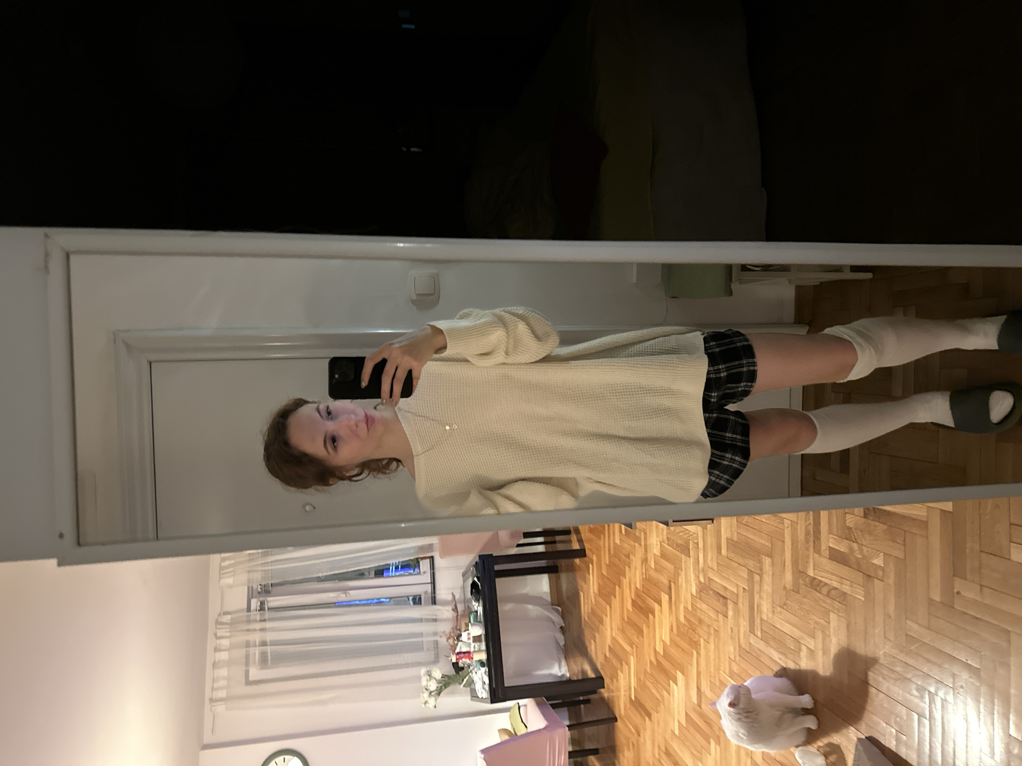A young woman is taking a mirror selfie. She is wearing a cream-colored sweater, a plaid skirt, and white knee-high socks with dark sandals. She is holding a smartphone up to her face, and her reflection shows a room with hardwood floors and a window with white curtains. A white cat sits on the floor. The room is in Novi Sad, Serbia.