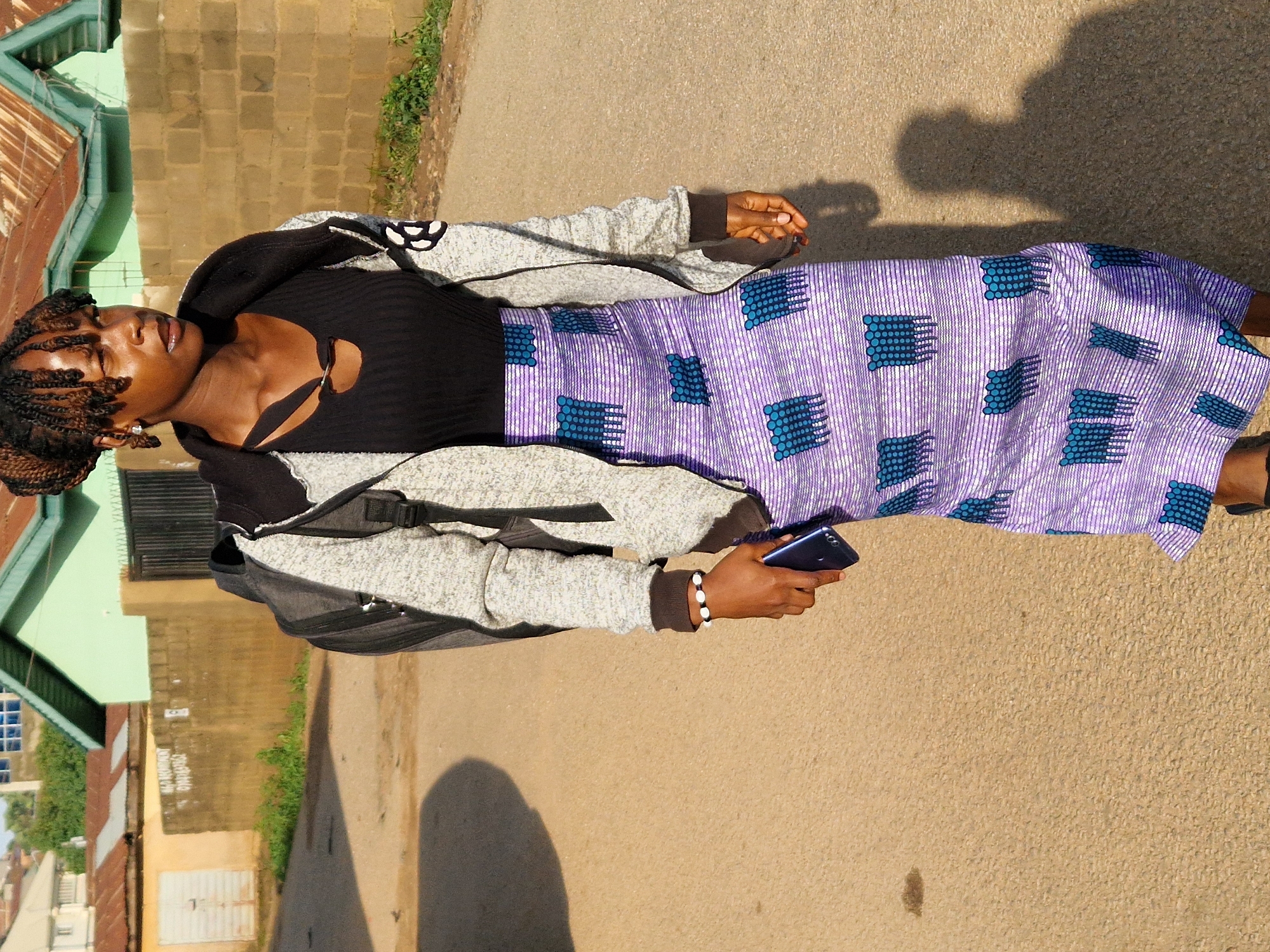 The image depicts a young woman standing on a paved road in Kabong, Nigeria. She is wearing a black ribbed top with a cutout detail, a grey hooded jacket, and a long purple skirt with a white and blue geometric pattern. A black backpack is on her shoulders. In her right hand, she holds a dark-colored smartphone. She is wearing a bracelet with white beads on her left wrist. The background shows a wall made of beige bricks, a metal gate, and buildings with green and red corrugated metal roofs. The ground is a light brown, asphalt-like surface, and a shadow of the woman is cast on it. The lighting suggests it is daytime, possibly late afternoon, with sunlight creating strong shadows.