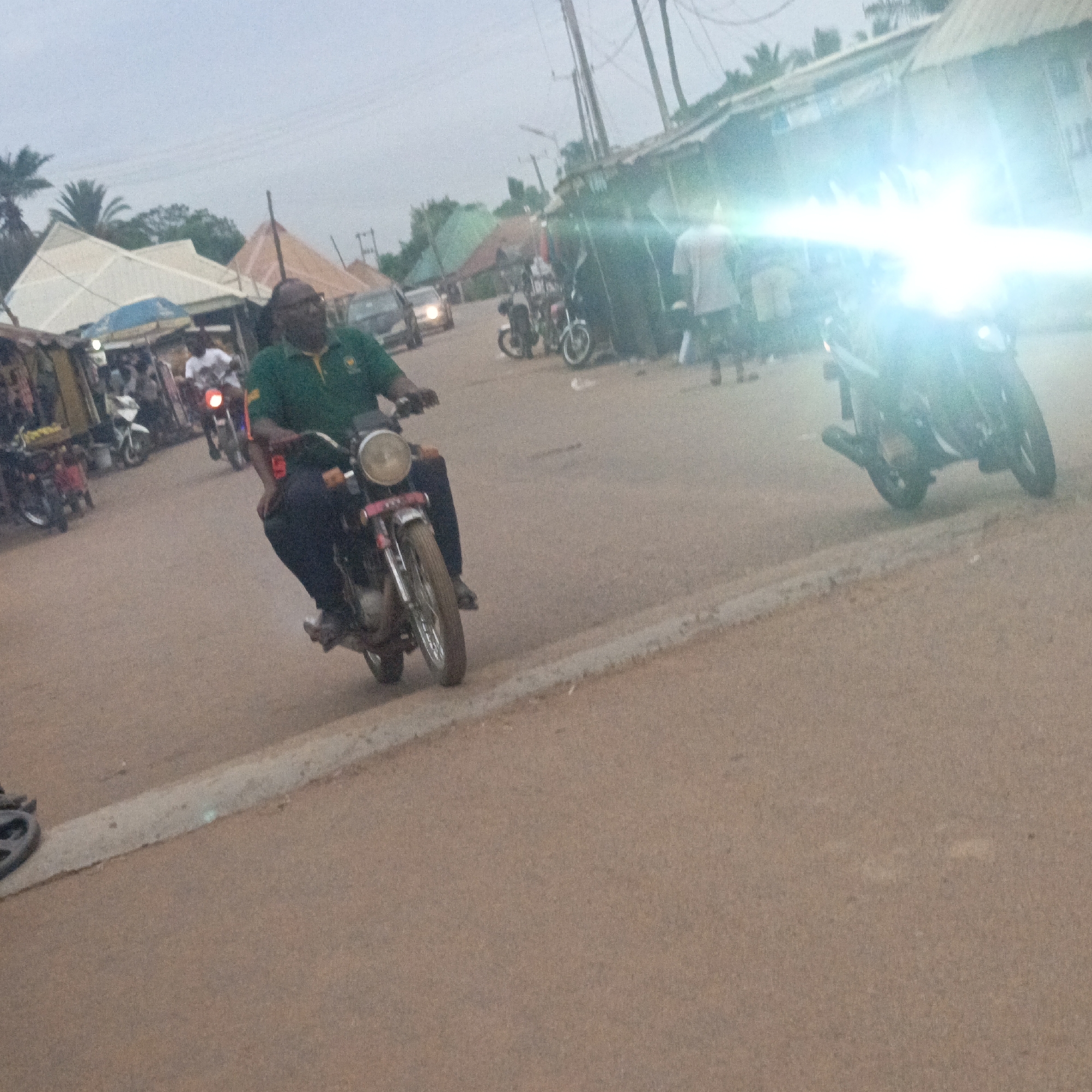 The image depicts a street scene in Akwanga, Nigeria. A man wearing a green shirt and dark trousers is riding a motorcycle in the foreground. Another motorcycle with a bright headlight is approaching in the distance. Along the roadside are shops and stalls. People are present near the shops. A car is visible further down the road. Palm trees are in the background.