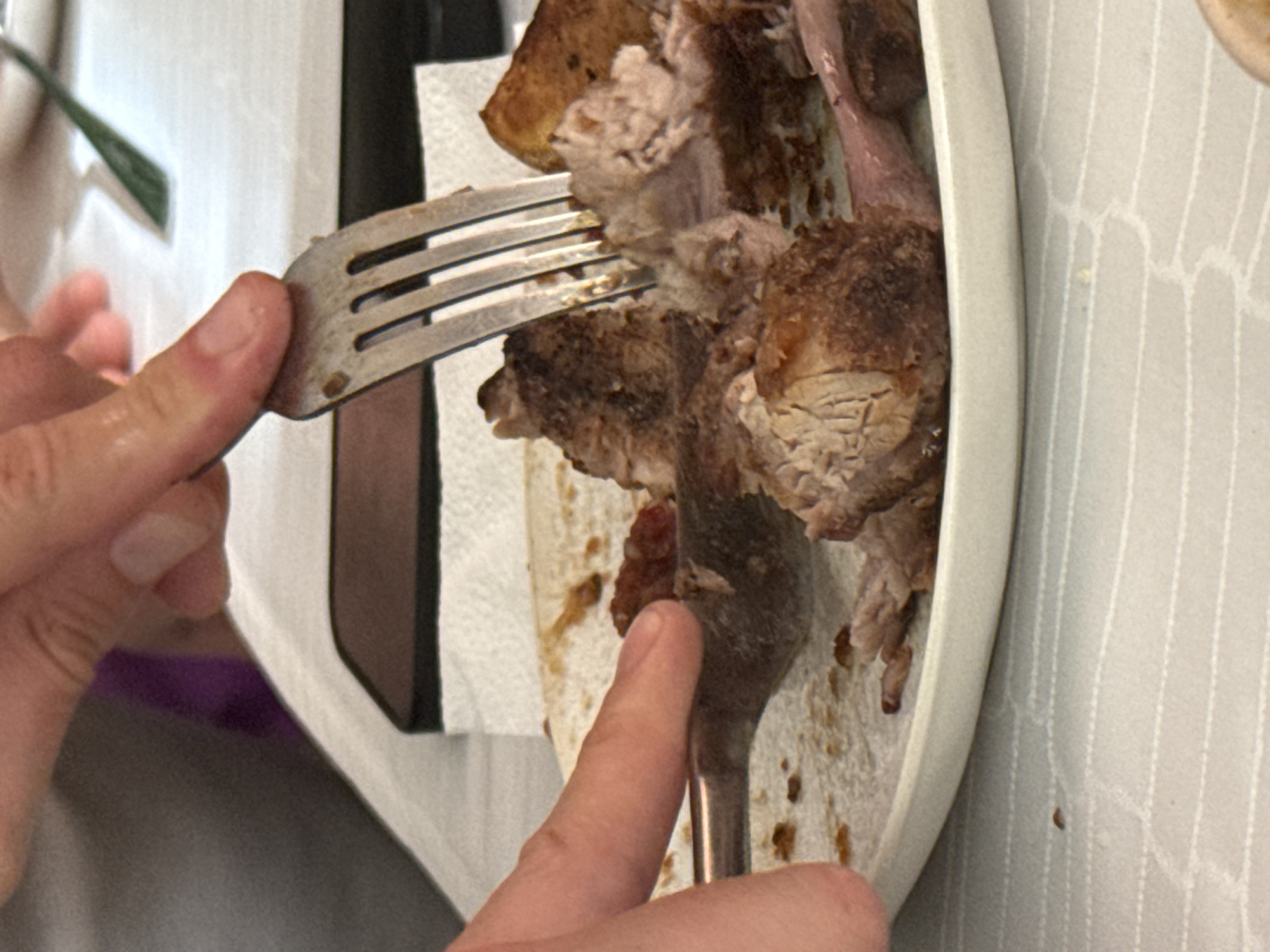 A close-up shot focuses on a person's hands using a fork to pull apart pieces of cooked meat on a white plate. The meat appears to be a roast, possibly pork or beef, with visible layers of muscle and fat. Some pieces are darker brown, indicating they are well-cooked or have a crust, while others are lighter pinkish-brown. A dark red sauce or condiment is visible on the plate near the meat. The plate rests on a white tablecloth with a subtle pattern. In the background, another fork with a dark handle is partially visible, as is a portion of a dark rectangular object, possibly a phone or tablet, lying flat. The overall scene suggests a meal is being prepared or consumed. The image does not provide any information that would allow for its location to be identified in Pavlohrad, Ukraine, or any other city.