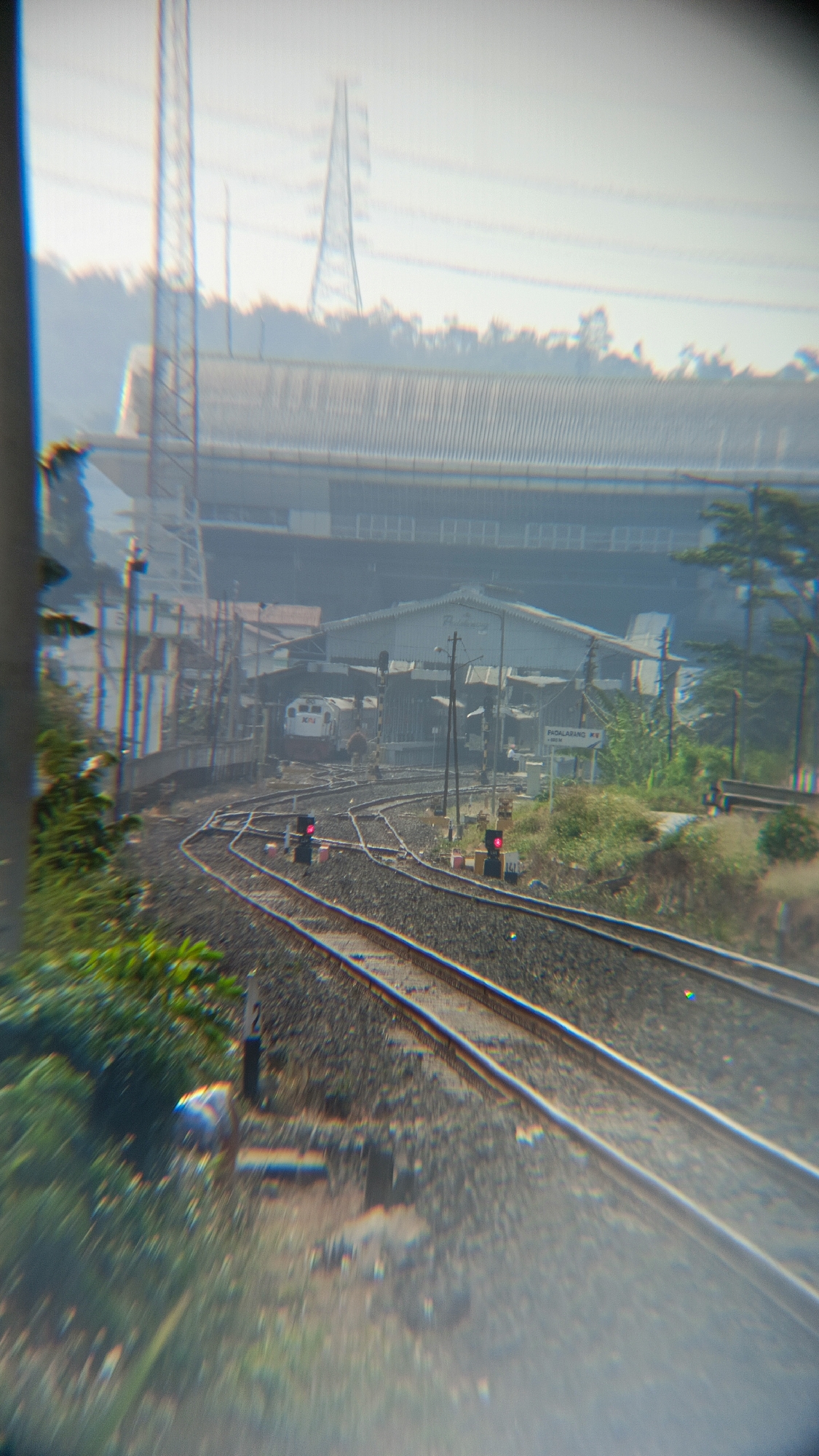 The image depicts a train station with multiple railway tracks. In the background, a large, elongated building with a corrugated roof and a section of elevated structure is visible, possibly a station hall or a maintenance facility. Adjacent to the building, there are several smaller structures and a train, partially obscured by the tracks. A sign in the distance reads "PADALARANG". Two sets of railway signals with red lights are positioned along the tracks. The foreground shows a dense cluster of green foliage. The overall environment appears to be an outdoor railway yard with elements of urban infrastructure and vegetation.