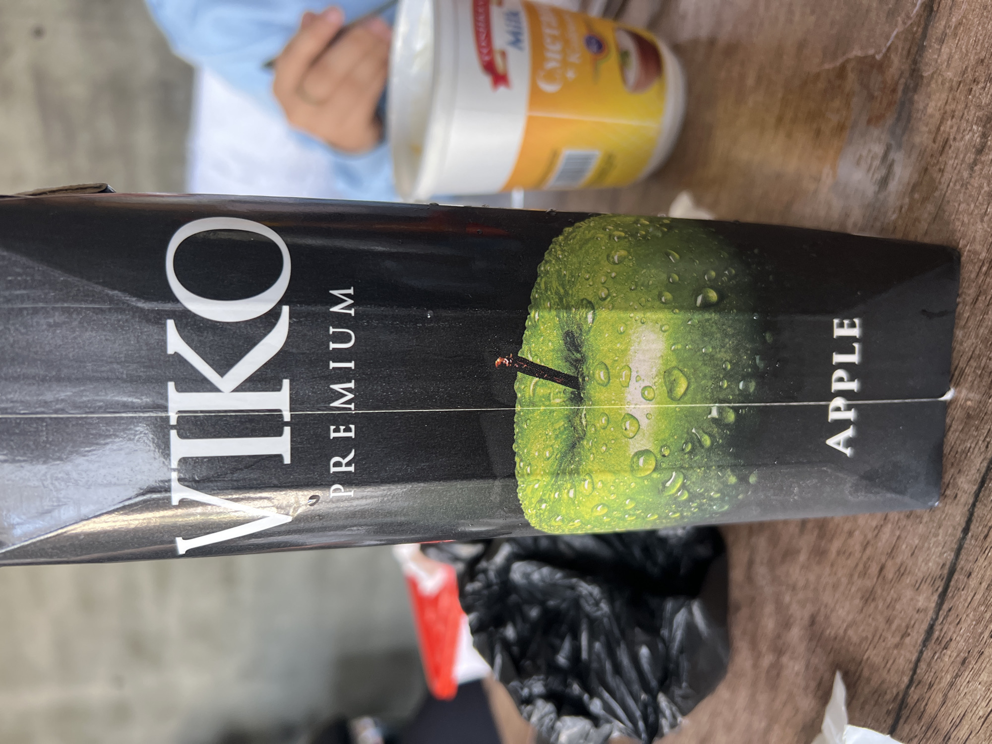 A close-up, eye-level shot shows the front of a black carton of VIKO PREMIUM apple juice. The carton has a glossy finish and features a realistic image of a dew-covered green apple. White text on the carton reads "VIKO PREMIUM" at the top and "APPLE" at the bottom. To the right of the carton, a partially visible white plastic tub with a yellow label containing Cyrillic text is present. In the background, out of focus, is a person's hand and arm, possibly reaching for something. The scene appears to be set on a wooden surface, suggesting a table. The overall impression is that of a casual setting, possibly a meal or snack time. The image does not contain any explicit information indicating its location in Shahrixon, Uzbekistan.
