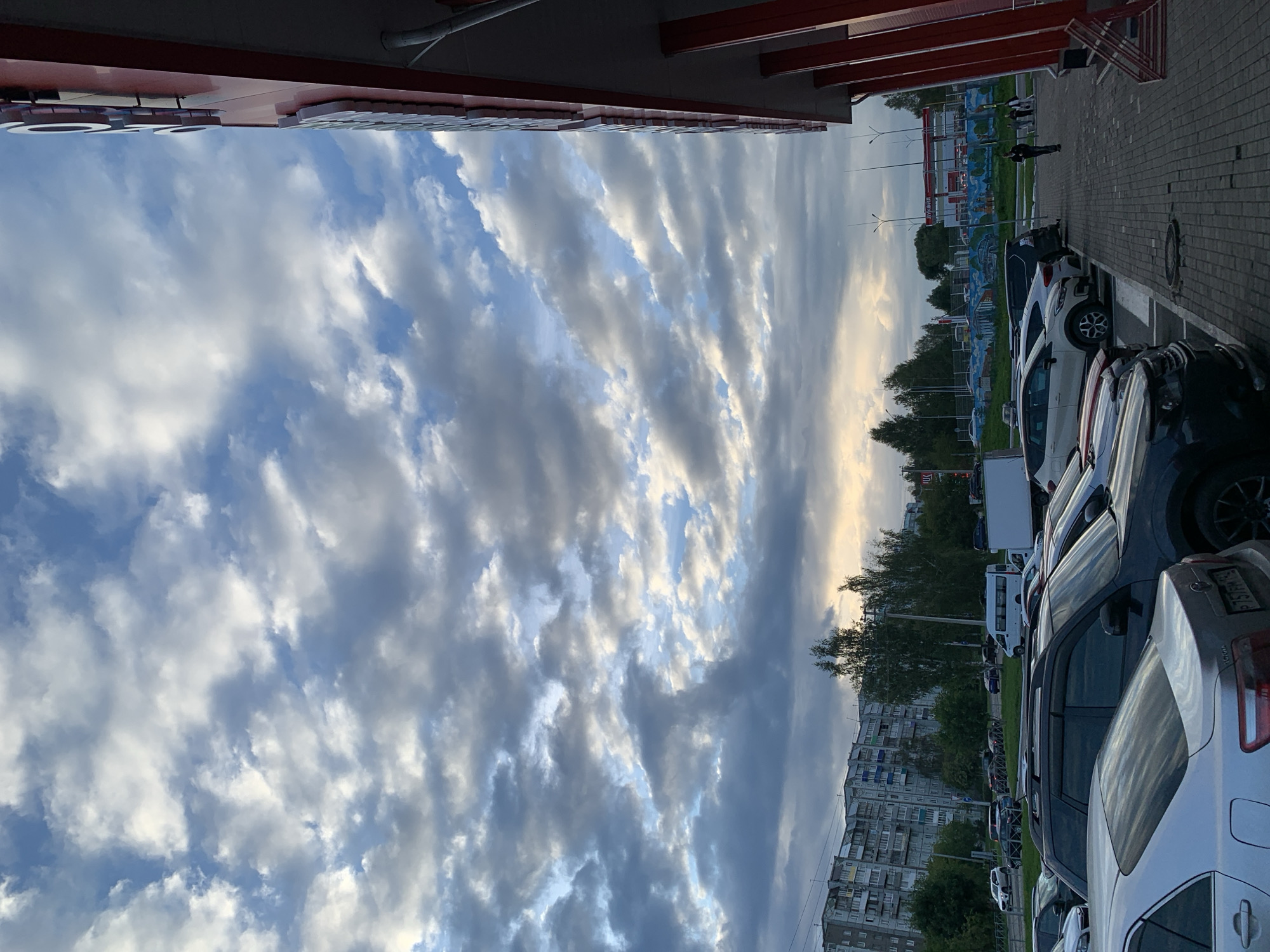 The image depicts a wide shot of a parking lot outside a commercial building, under a sky filled with scattered clouds. The perspective is from ground level, looking upwards and towards the right. On the right side of the frame, a portion of a building with a red facade and white lettering is visible, extending towards the top of the image. The building appears to be a commercial establishment.
In the foreground and midground, numerous cars are parked in rows. The vehicles vary in type and color, including sedans, SUVs, and a white van. The parking lot is paved with asphalt and marked with white parking lines. A paved walkway runs alongside the building on the right.
In the background, a row of apartment buildings with a grey facade is visible on the left, suggesting an urban or suburban setting. Trees are interspersed between the buildings and along the edge of the parking lot. In the far background, a white truck is parked, and further beyond, there appears to be a fuel station with red signage. A fence with a mural is also visible in the background.
Several people are present in the scene. Two individuals are walking on the paved walkway near the building. Another person is walking in the distance, towards the right side of the frame. A group of people are standing together further down the walkway.
The sky is a prominent feature, with a mix of white and dark grey clouds against patches of blue sky, suggesting it is daytime, possibly late afternoon or early evening, given the lighting. The overall impression is a typical scene in a commercial area of a Russian city. The image is likely taken in Prokopyevsk, Russia, given the architectural style of the buildings and the presence of Cyrillic signage in the background.