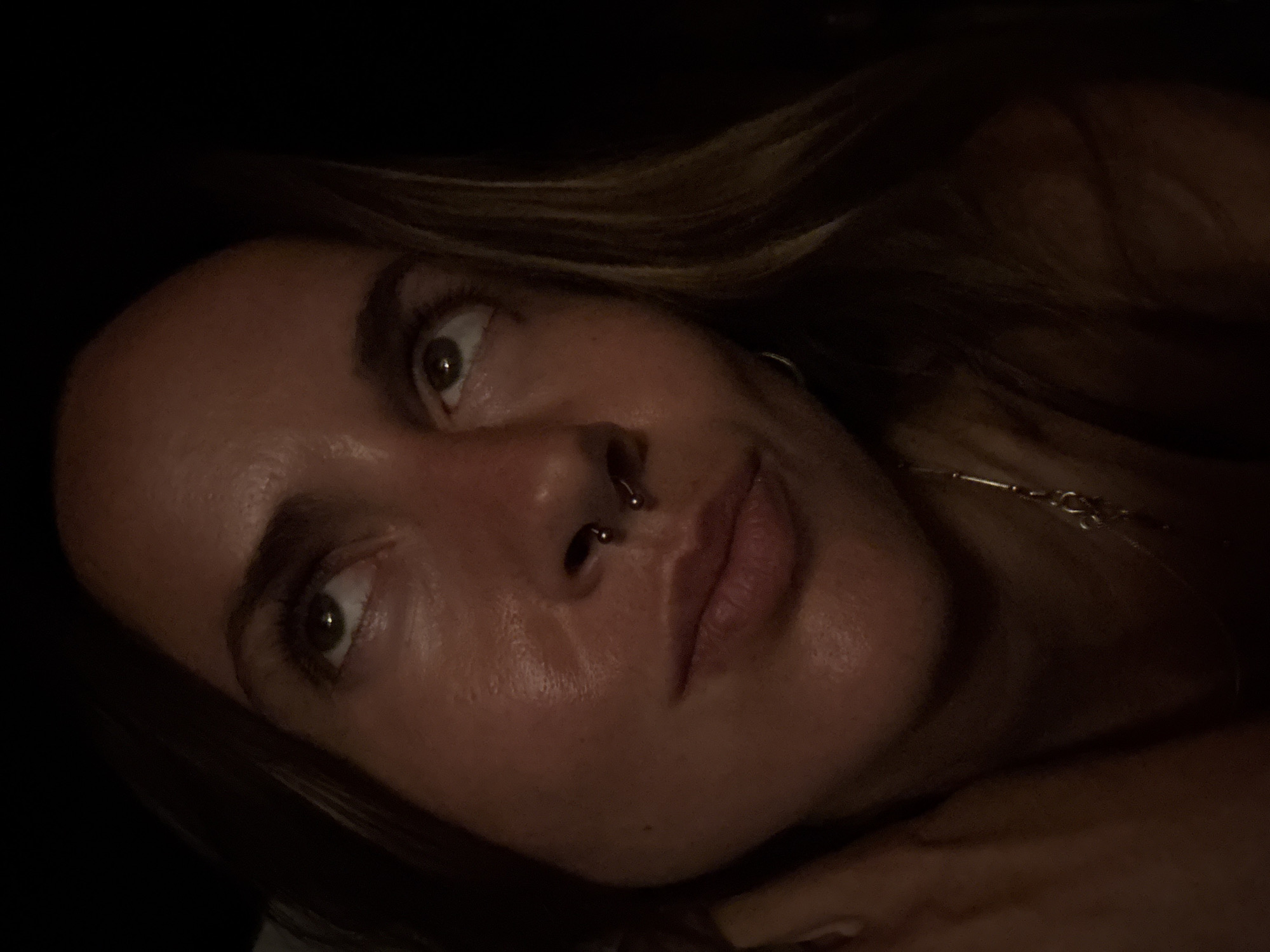 A woman is looking up and to the left, her face illuminated by an unseen light source from above and to her right. She has brown hair, a septum piercing, and is wearing a necklace. Her skin appears to be slightly oily. The background is dark and indistinct. The image appears to be a close-up portrait taken in a low-light environment.