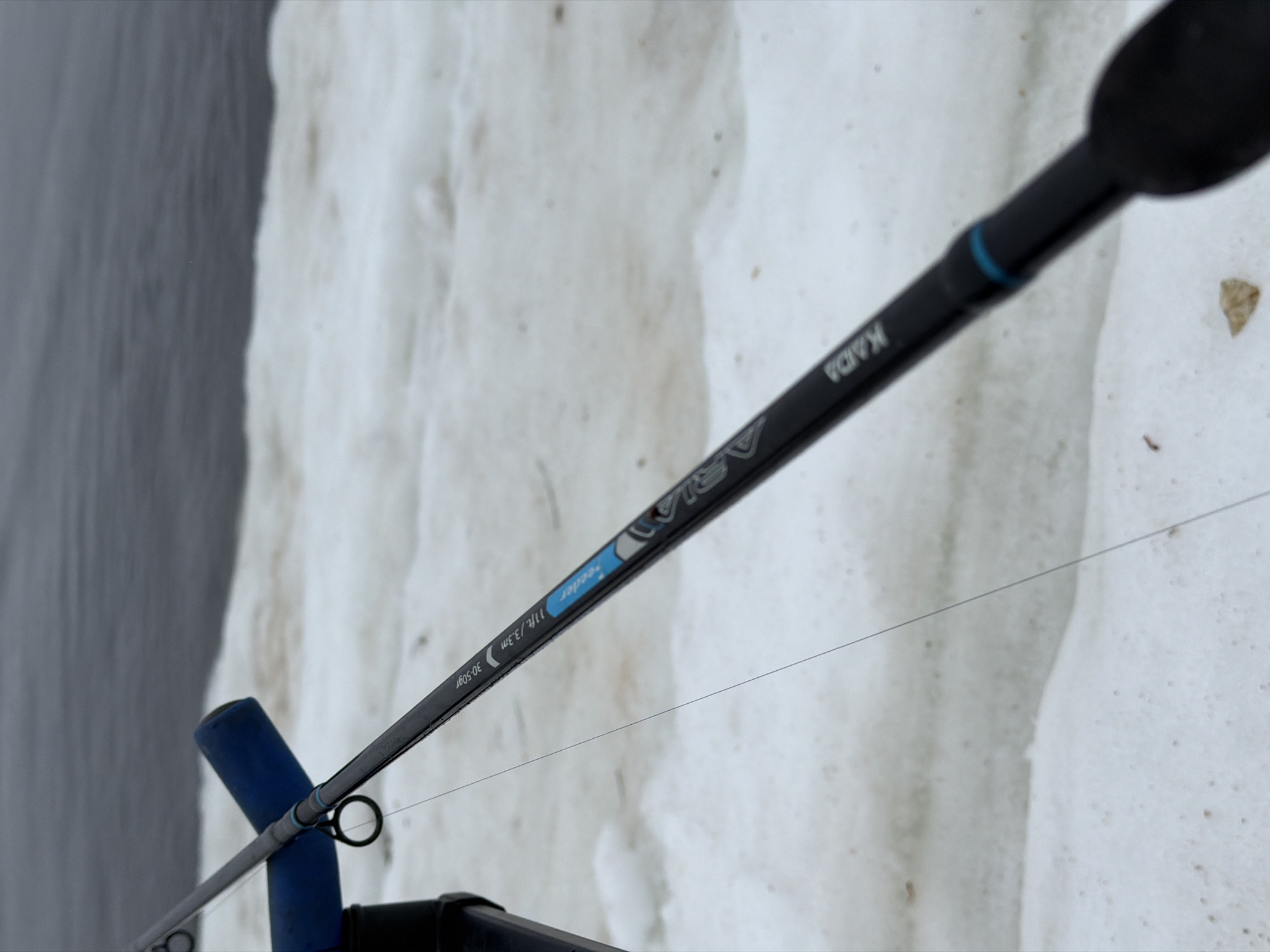 The image shows a close-up of a fishing rod, identified by the brand "KAIDA" and model "Aria Feeder", with the specifications "11ft/3.3m" and "30-50gr" visible on the rod's surface. A fishing line is threaded through the rod guides. The rod is positioned on a support, and a portion of a body of water is visible on the left side of the frame, contrasted with a snowy or icy bank on the right. Based on prior information, this scene is located in Chaikovsky, Russia.