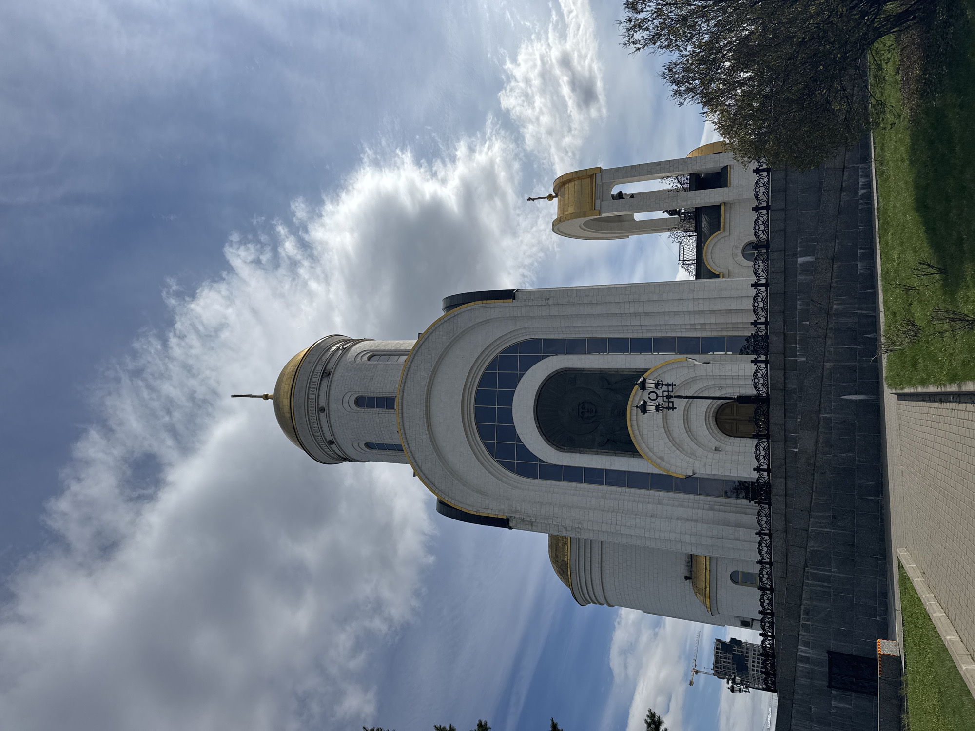 The image depicts the Victory Park in Moscow, Russia. In particular, the image shows the Church of St. George the Victorious, which is located in this park. The church is white with gold domes. There is a blue sky with some white clouds. A tree and a sidewalk are visible. In the background is some modern construction.