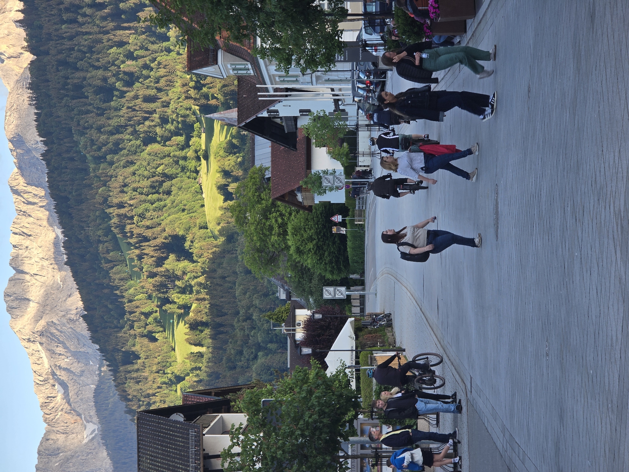 The image depicts a vibrant street scene in Garmisch-Partenkirchen, Germany, framed by the majestic Alps. In the background, towering, rugged mountains, likely part of the Wetterstein range, dominate the horizon. These peaks exhibit a light, rocky texture with visible patches of snow or ice in shaded crevices, and they are brightly illuminated by direct sunlight, suggesting either early morning or late afternoon. Below the mountains, a dense, dark green coniferous forest covers undulating hills. Several patches of bright green meadowland are interspersed within the forest, also catching the sunlight. The midground features the town's architecture, characterized by traditional Bavarian-style buildings with pitched roofs, some featuring dark tiles and others reddish-brown. Many buildings have light-colored facades, dark-colored shutters, and balconies. On the right, a multi-story building displays a sign that partially reads "PARK" and "Hauptstrasse." A conical spire, possibly belonging to a church or chapel, is visible above some roofs on the right side of the street. The paved street, possibly cobbled, gently curves to the right and appears to be a main thoroughfare. On the right side of the street, there's a sidewalk and several parked cars further down. Traffic signs, including a "20 km/h" speed limit and a no-passing zone, are visible on the right. A white tent structure with "PAULANER" printed on it indicates an outdoor seating area. Planters with purple and pink flowers are visible on the far right. In the foreground, the grey-toned paved street is populated by several individuals. On the far left, a person in a blue jacket and white shorts stands facing right, next to a man in a yellow shirt and dark pants who is also facing right. Another man in a dark jacket and blue jeans stands with his back to the viewer, talking to a blonde-haired woman. Adjacent to this group, a man on a black electric bicycle, wearing a dark jacket and blue helmet, rides away from the viewer. Further into the foreground, a woman in a light-colored t-shirt and dark jeans, carrying a black backpack, walks towards the left, partially obscured. Crossing the street from right to left, a woman in a horizontal striped shirt and blue jeans carries a red bag. Behind her, a cyclist in a black and white top with a helmet rides an e-bike. To their right, two women walk side-by-side from right to left. One wears a dark denim jacket and dark pants,