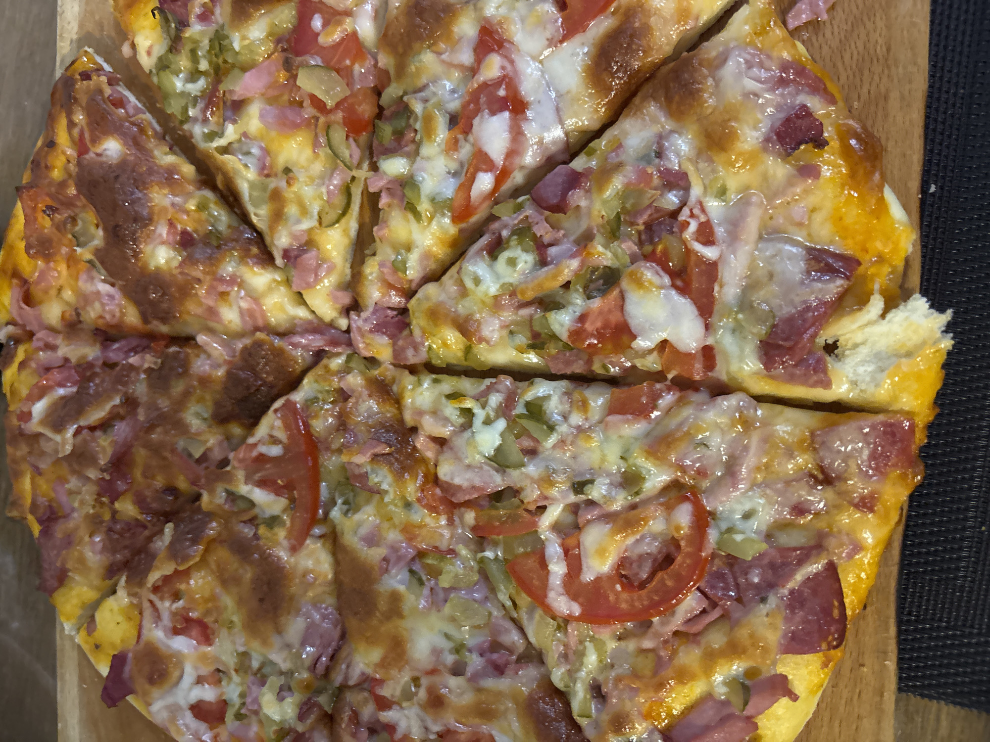 The image shows a pizza cut into slices on a wooden surface. The pizza toppings include cheese, tomatoes, ham, and green pickles. The photo was taken in Chaikovsky, Russia.