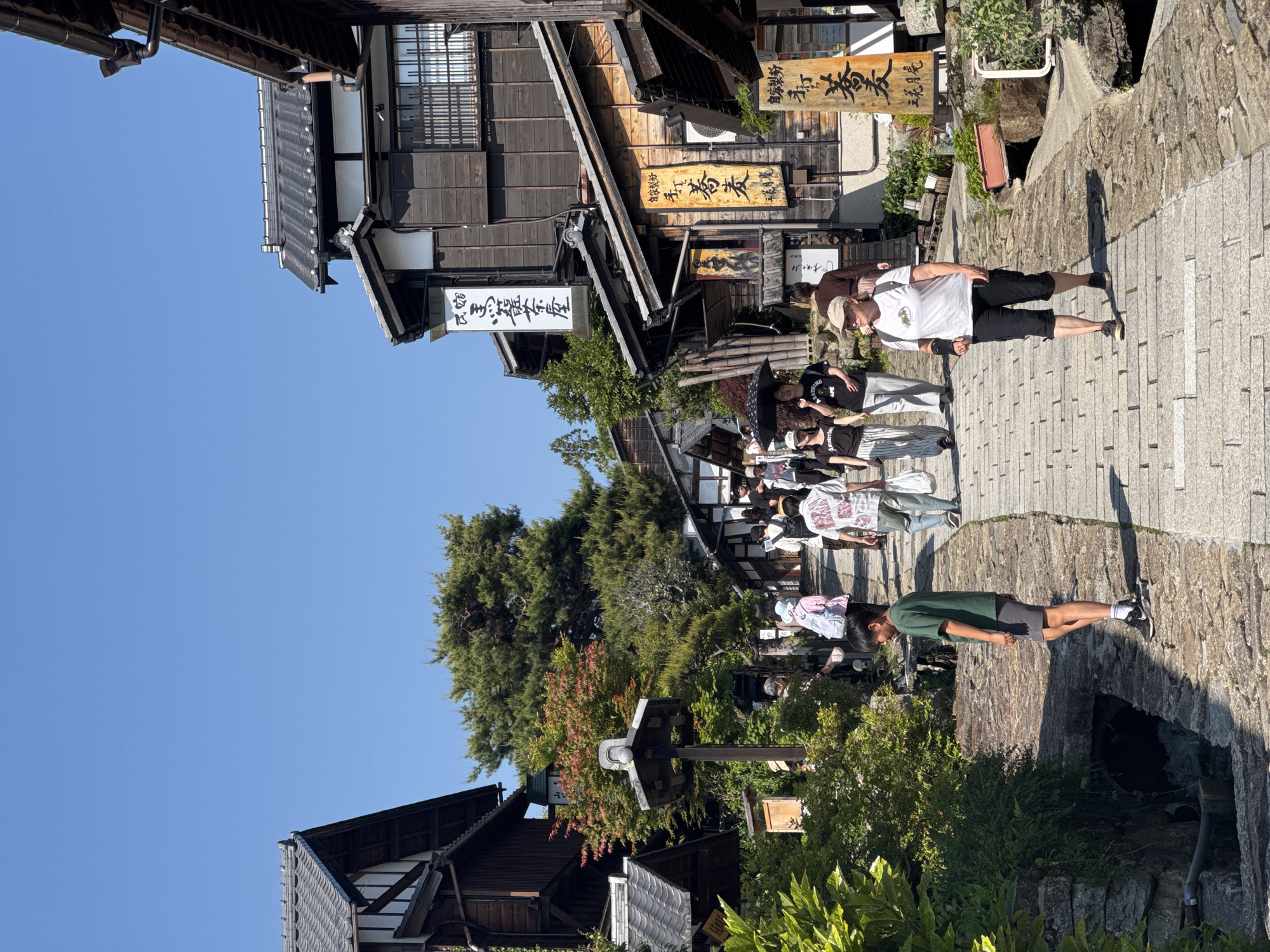 This image shows a street in the city of Nakatsugawa, Japan. The street is paved with cobblestones and lined with traditional Japanese buildings. There are several people walking along the street, including one man wearing a white t-shirt and black shorts and a person wearing a green t-shirt and grey shorts. The buildings are made of wood and have traditional Japanese roofs. There are also some trees and plants along the street. The sky is blue and clear.
