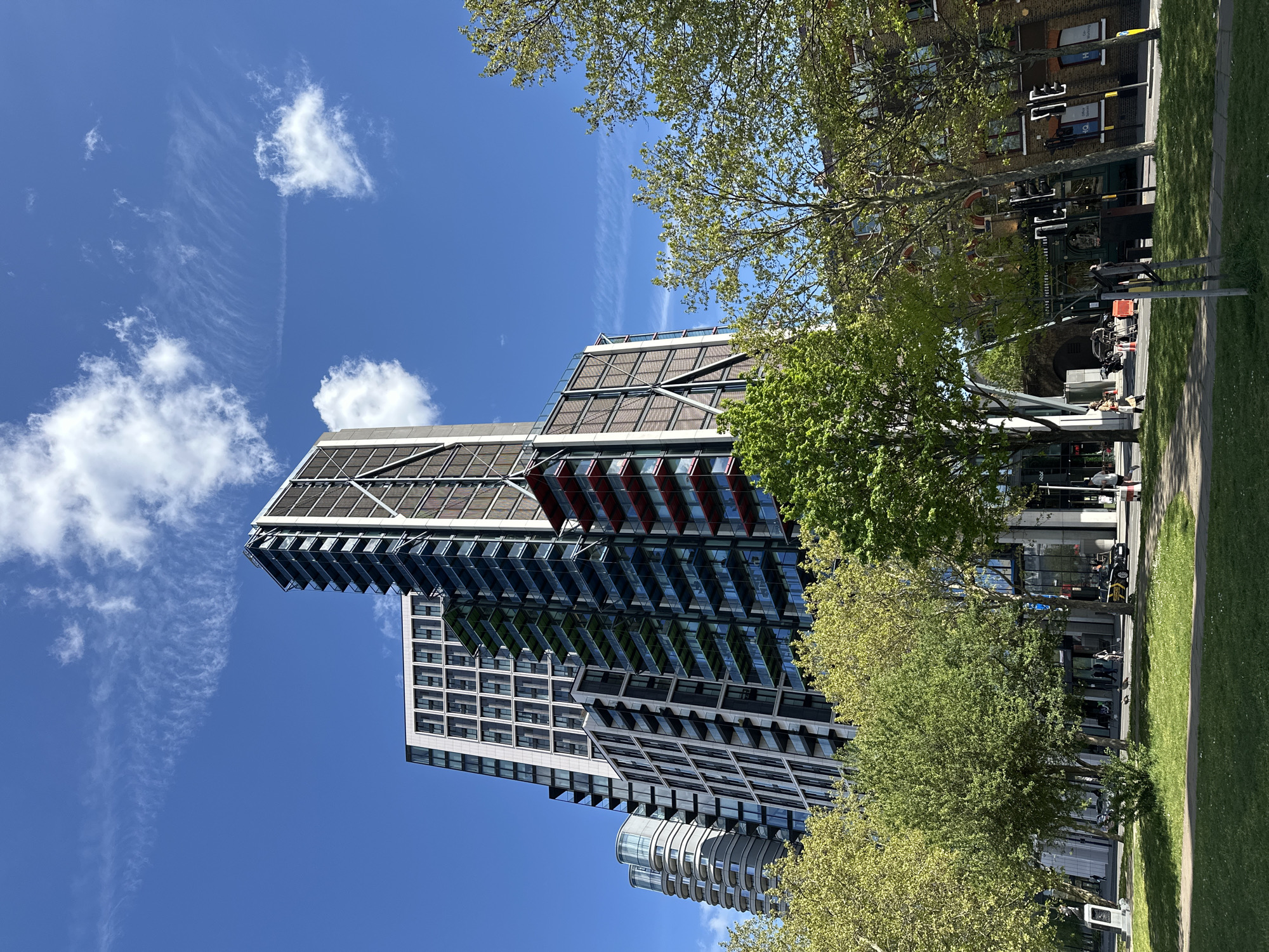 The image depicts a modern architectural landscape in London, United Kingdom. A high-rise building dominates the scene, showcasing a sleek, contemporary design with a mix of glass and metal structures. The building appears to be residential, with multiple balconies.
The sky is bright blue with scattered white clouds. Trees with green foliage frame the building, creating a natural contrast to the urban environment.
On the ground level, a public space is visible, featuring a paved walkway and grassy areas. There are people walking along the path. A road with cars and pedestrian crossing signals is present. Behind the trees are office buildings.