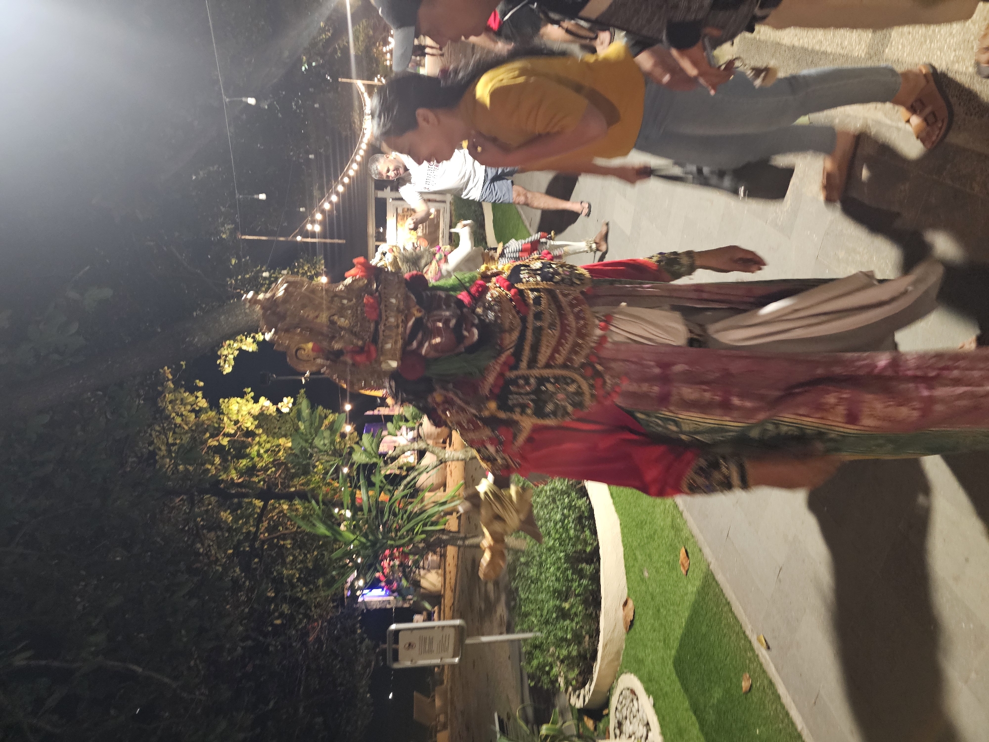 Here's a forensic description of the image:
**Scene:** An outdoor setting at night. Overhead string lights illuminate a pathway alongside garden vegetation.
**Subject:** A person in ornate Balinese costume and headdress. The costume is predominantly red and gold.
**People:** Several individuals are also present. One woman wears a yellow shirt and light blue jeans, and appears to be looking at the costumed person. Another man is wearing a white shirt and blue shorts.
**Objects:** Signage is visible in the foreground near the garden.
**Location:** Potentially Denpasar, Indonesia, based on the cultural context of the Balinese costume and headdress.