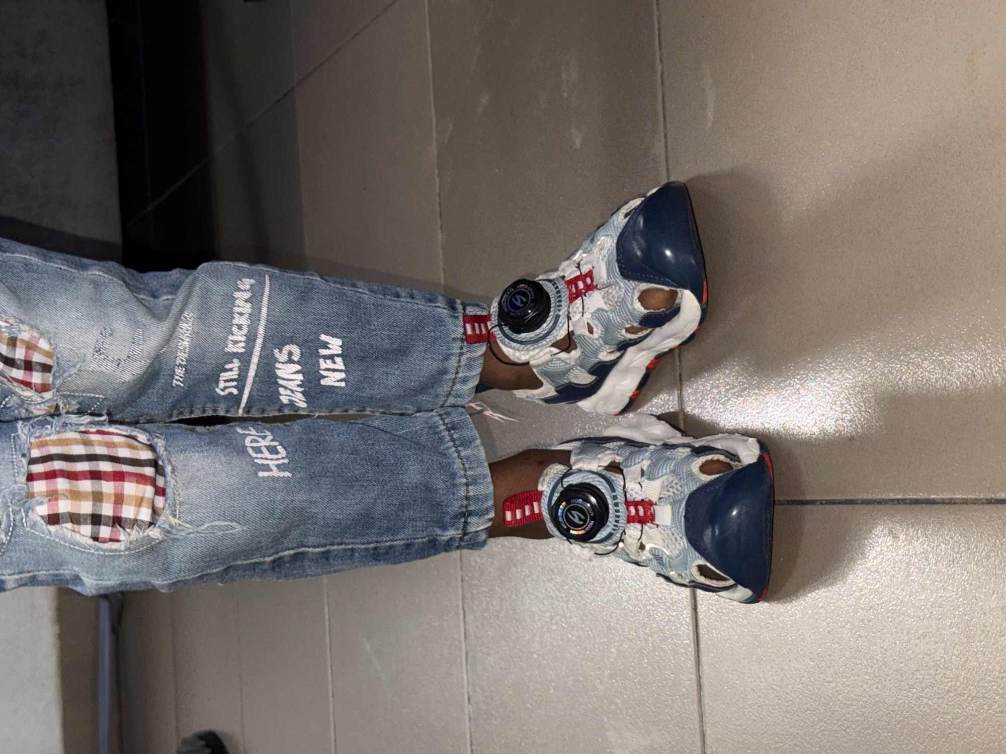 This low-angle, vertical shot captures the lower half of a person standing or sitting, focusing on their footwear and distressed jeans. The scene is set indoors on a light beige or grey tiled floor, likely in Ado, Nigeria, given the provided context.
The person is wearing light blue denim jeans that are heavily distressed. The left leg features a large torn area on the thigh, revealing a red, black, and beige plaid patch underneath. Another smaller tear is visible on the knee. White stenciled text is printed on the jeans; on the left leg, it reads "THE ORIGINAL", "STILL KICKIN'", "JEANS NEW", and "HERE". The word "HERE" is also visible on the lower part of the right leg.
On their feet, the person wears chunky athletic sneakers. These shoes incorporate shades of light blue, white, red, and dark blue (navy). A prominent feature is a circular black dial on the side of each shoe, which appears to be part of a lacing or tightening system, similar to a Boa Fit system. The dial has a logo, possibly stylized 'SB', and the word "HOOLIGAN" visible around its edge. Red webbing straps and white mesh-like material with cutouts contribute to the shoe's intricate design. The overall impression is casual and fashionable. No specific activity is depicted beyond the display of clothing and footwear.