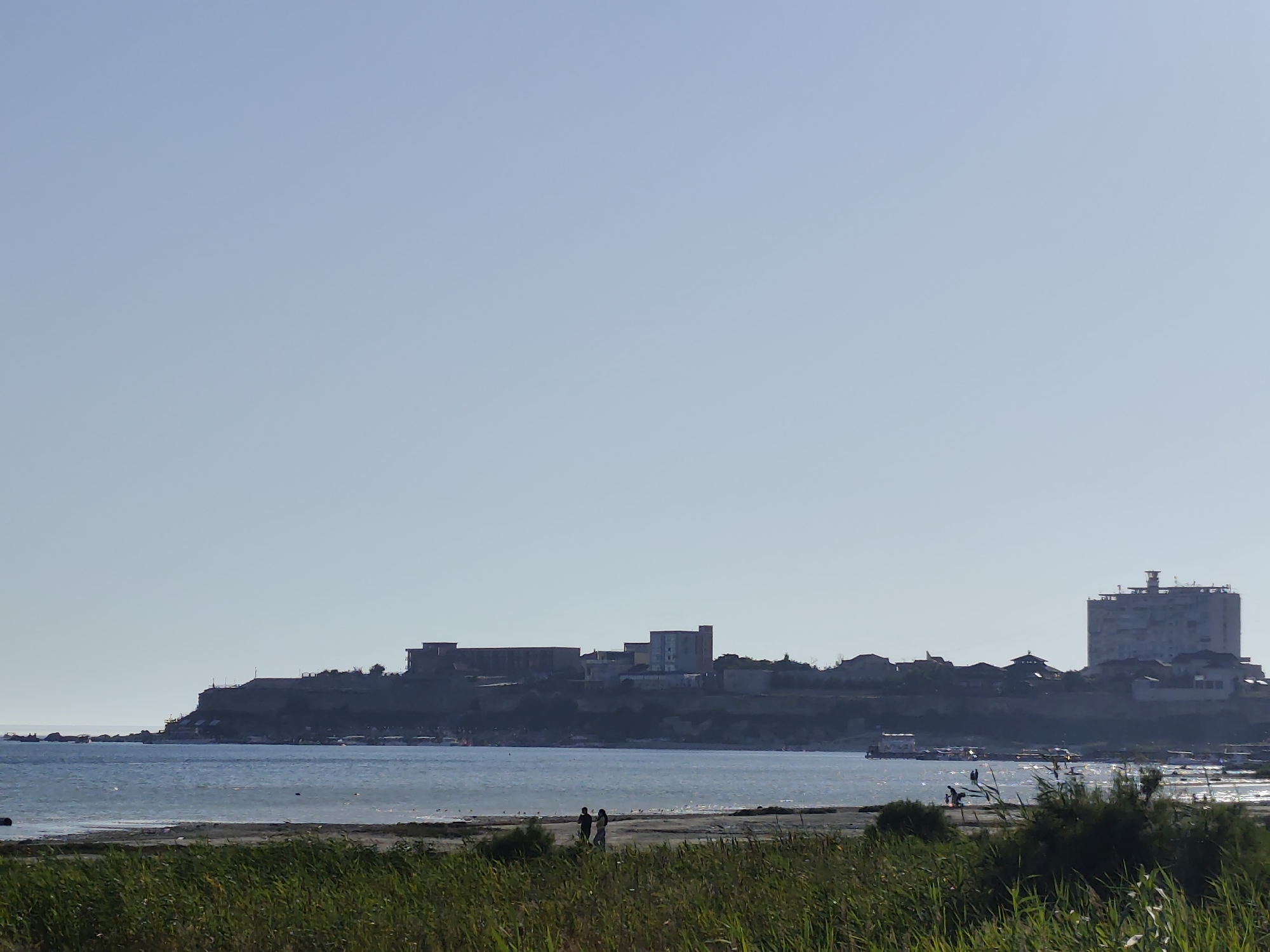The image shows a coastal landscape with a city on a bluff overlooking a body of water. In the foreground, there is tall, dry grass. The middle ground features a sandy shore with two silhouetted figures walking, and several scattered birds. The water is calm and reflects the light. The background displays a city with various buildings, including a prominent, large, light-colored building on the right side of the frame. Boats are visible in the water near the shore. The sky is clear and light blue. This scene depicts a view of the Caspian Sea coastline near Aktau, Kazakhstan, possibly showing the area of the "Mayak" settlement and the city's promenade.