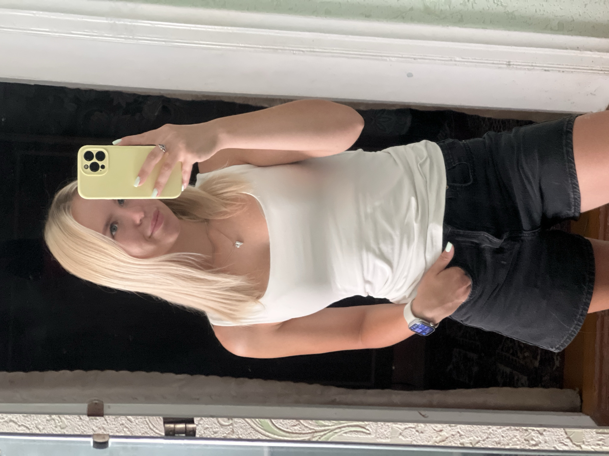 A young Caucasian woman with blonde hair is taking a mirror selfie. She is wearing a white tank top and black denim shorts. She has a watch on her left wrist and a ring on her right index finger. She is holding a yellow smartphone in her right hand, which is angled to capture the reflection. The background appears to be an interior space, possibly a hallway or doorway, with a dark opening behind her and a textured wall to her left. The lighting suggests it is daytime.
