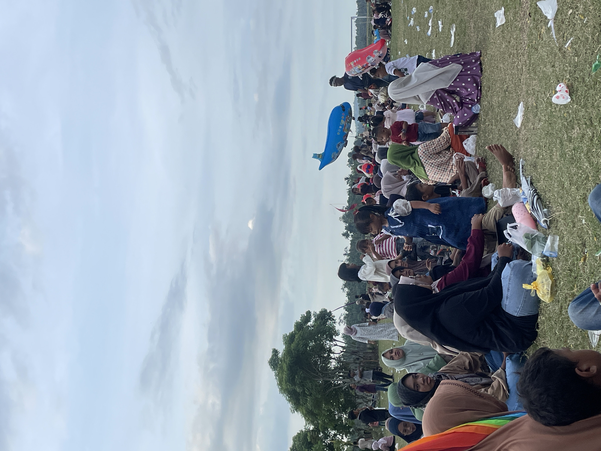 A crowd of people, predominantly women and children wearing hijabs and modest clothing, are gathered on a grassy field, likely in Geuleumpang Geuleudieng, Indonesia. They are seated on the ground, some interacting with each other, while others are looking around or observing activities. Several children are visible, some standing and playing, while others are seated with adults. In the background, trees and a fence are discernible. The sky above is overcast with scattered clouds. Two large, inflatable balloons, one shaped like a blue airplane and another like a red balloon, are suspended in the air. Litter, including plastic bags and wrappers, is scattered across the grass, suggesting a public event or gathering. The overall atmosphere appears to be casual and community-oriented.