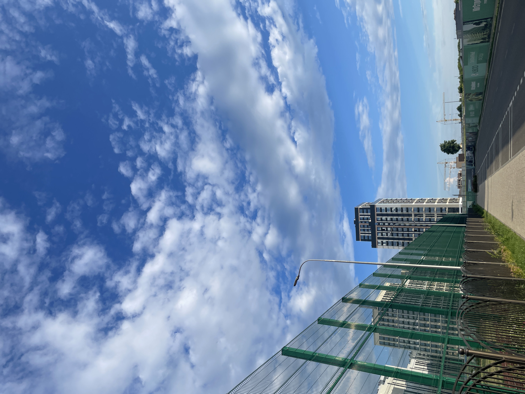 The image depicts an outdoor urban scene with a clear blue sky filled with scattered white clouds. In the foreground, a paved walkway runs parallel to a tall green wire mesh fence, which is topped with a decorative black metal fence. Beyond the fence, a modern high-rise residential building with a dark facade and numerous windows stands tall. To the left of this building, other residential buildings are visible. Further in the distance, construction cranes are apparent, suggesting ongoing development. The ground adjacent to the walkway is covered with grass. The scene appears to be a residential development area in Stavropol, Russia, given the construction activity and modern apartment buildings. There are no people visible in the image.