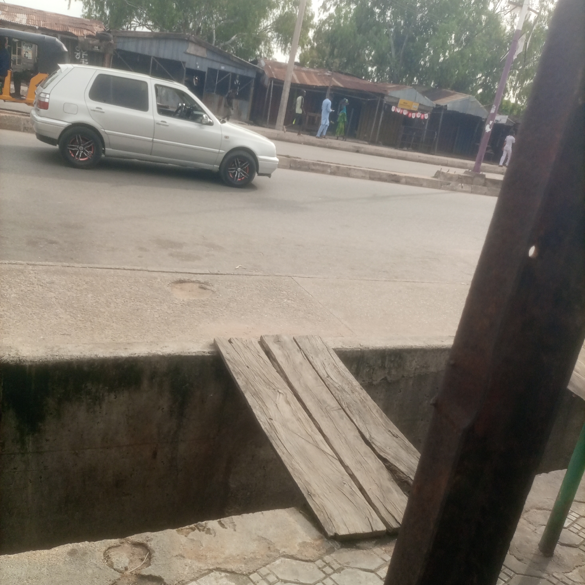 The image depicts a street scene in Jalingo, Nigeria, characterized by its open road, adjacent structures, and a notable disparity in pavement quality. A silver Volkswagen Golf hatchback is parked or moving on the paved road. Several individuals are visible, including one in light blue attire and another in green, walking along the sidewalk. Behind them, various small shops or stalls with corrugated metal roofs line the street. The foreground features a concrete structure with wooden planks laid across a gap, possibly serving as a makeshift bridge over a drainage area. The overall environment suggests a busy urban setting with a mix of infrastructure and commercial activity.