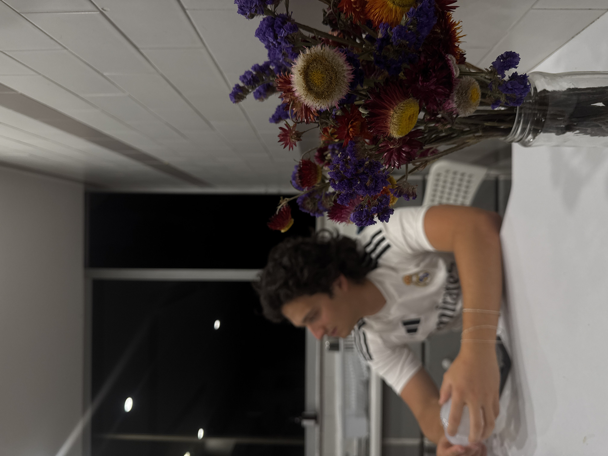 A vertical shot displays a person in a white Real Madrid jersey bending over a white surface, possibly a table, with a bouquet of colorful dried flowers in a glass vase positioned above them. The person has curly dark hair and their hand is near a clear plastic container. In the background, there's a window showing a dark, possibly nighttime, scene with some light sources visible. The setting suggests an indoor environment with a white tiled ceiling. The location is likely Mexico City, Mexico, given the request.