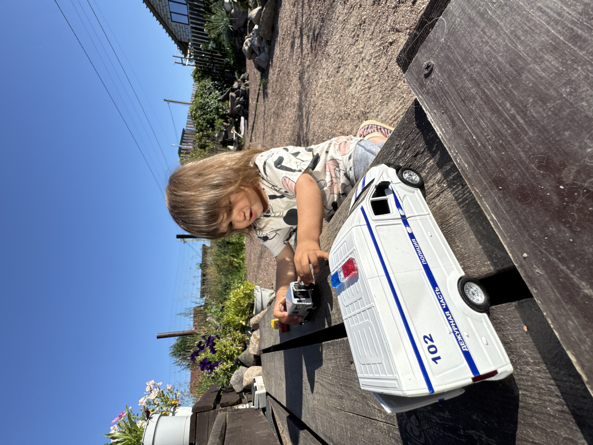A young child with long, brown hair is playing with toy vehicles on a weathered wooden table outdoors. The child is wearing a light-colored t-shirt with a pattern and grey shorts. In the foreground, a white toy police van with blue stripes and flashing red and blue lights is prominent. The van has the number "102" and Russian text "ДЕЖУРНАЯ ЧАСТЬ" (Duty Part) and "ПОЛИЦИЯ" (Police) printed on its side. Behind it, two smaller toy cars, one red and one yellow, are also present on the table. The background suggests a rural or suburban setting with a clear blue sky, green foliage, a fence, and a house. The ground appears to be covered in gravel. The image was taken in the city of Kempelevo, Russia.