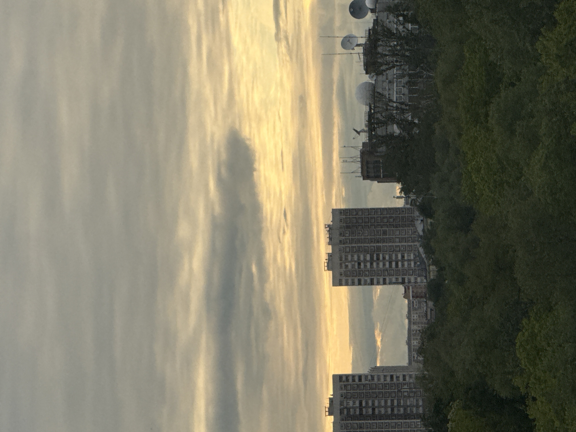 The image shows a view of Khimki, Russia, at dusk. The sky is filled with clouds, illuminated with a yellow and grey light. Several high-rise apartment buildings with regularly spaced windows are visible, partially obscured by trees on the right side of the frame. On one building in the distance, there is a series of satellite dishes on top.