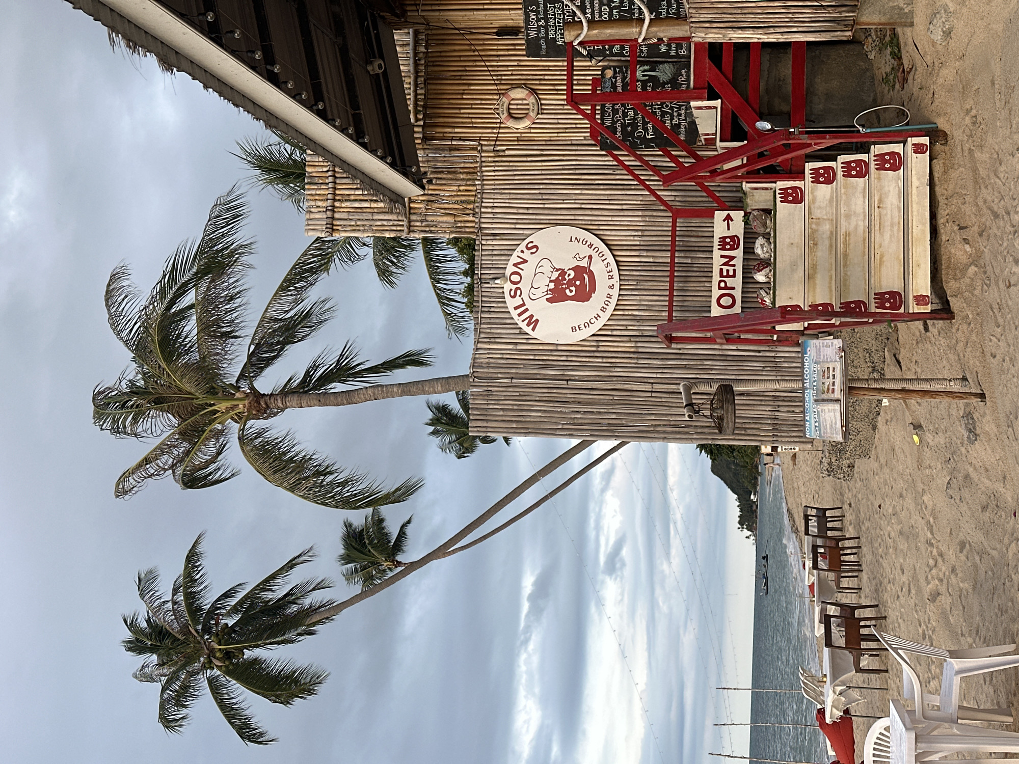 This is a vertical, eye-level shot of Wilson's Beach Bar & Restaurant in Baan Tai, Thailand. The building is constructed with bamboo, and palm trees with coconuts are visible. The sky is overcast with a cloudy, grey appearance, suggesting it might be a cloudy day or late afternoon.
The establishment appears to be open, indicated by a prominent "OPEN" sign with an arrow. A red staircase leads up to the building, with white steps decorated with a red cartoon face. A life preserver is mounted on the bamboo wall. Menu boards with handwritten items like "BREAKFAST," "APPETIZERS," "Skewer," "Thai F," "Danish," and "Soft Drinks" are visible. Further text mentions "NON-ALCOHOL ALCOHOL FISH & SALAD."
The restaurant is situated on a sandy beach overlooking the ocean. Several tables with white tablecloths and chairs are set up on the sand, facing the water, suggesting a dining or seating area for patrons. The presence of the tables and chairs indicates that customers are likely present or expected. The overall atmosphere appears relaxed and casual, typical of a beachfront establishment.