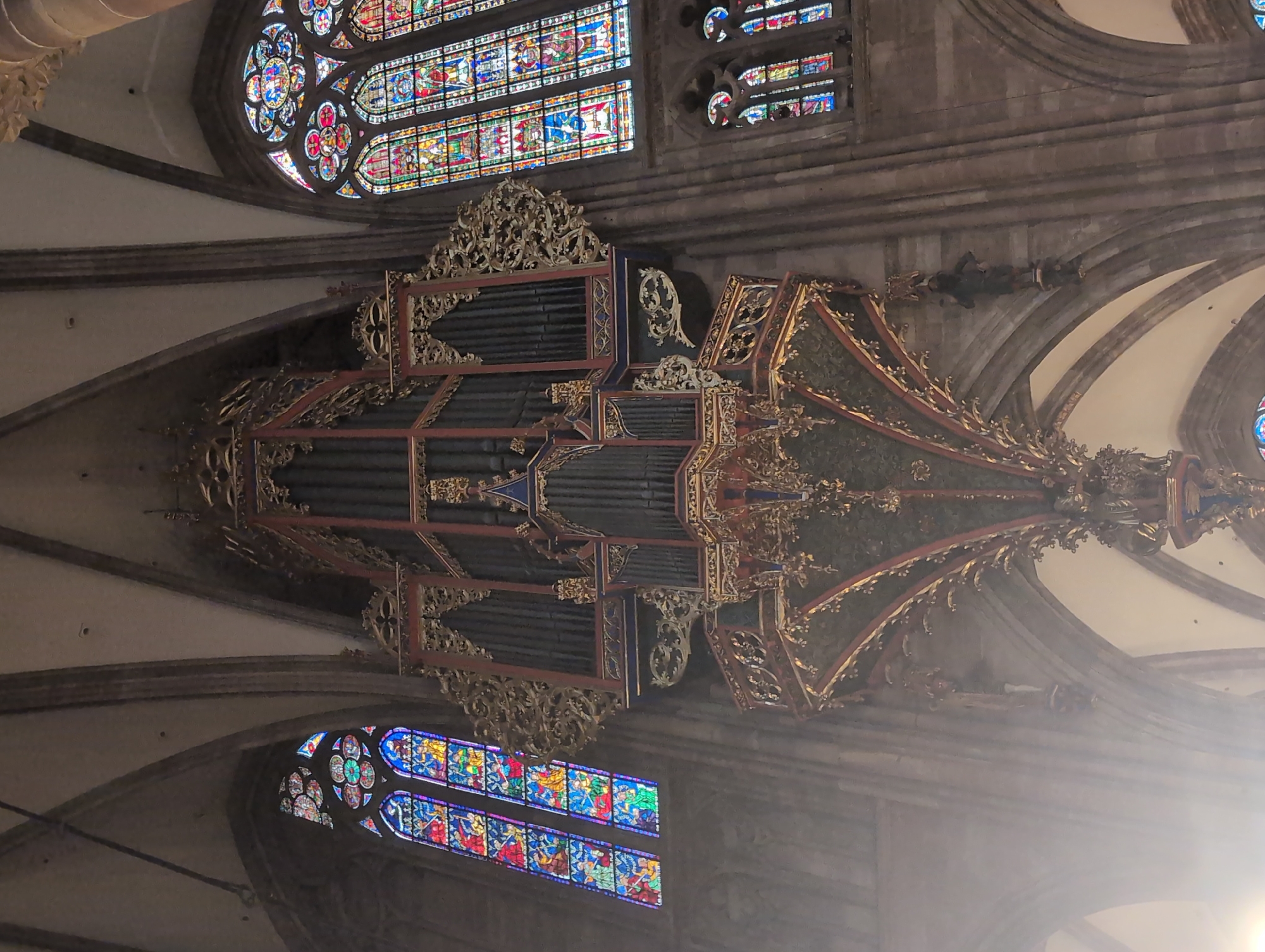 The image shows the interior of a Gothic cathedral, likely Strasbourg Cathedral, in Strasbourg, France. A large pipe organ, ornately decorated in gold and dark colors, dominates the center of the frame, suspended from the ceiling. The organ features numerous pipes and intricate carvings. To the left and right of the organ are tall, arched stained-glass windows filled with vibrant colors and detailed depictions. The architecture of the cathedral, with its high vaulted ceilings and stone columns, is visible. Light streams in, particularly on the left side, creating a hazy effect. Small, dark figures are visible affixed to the architectural elements on the right side of the organ, appearing to be decorative sculptures.