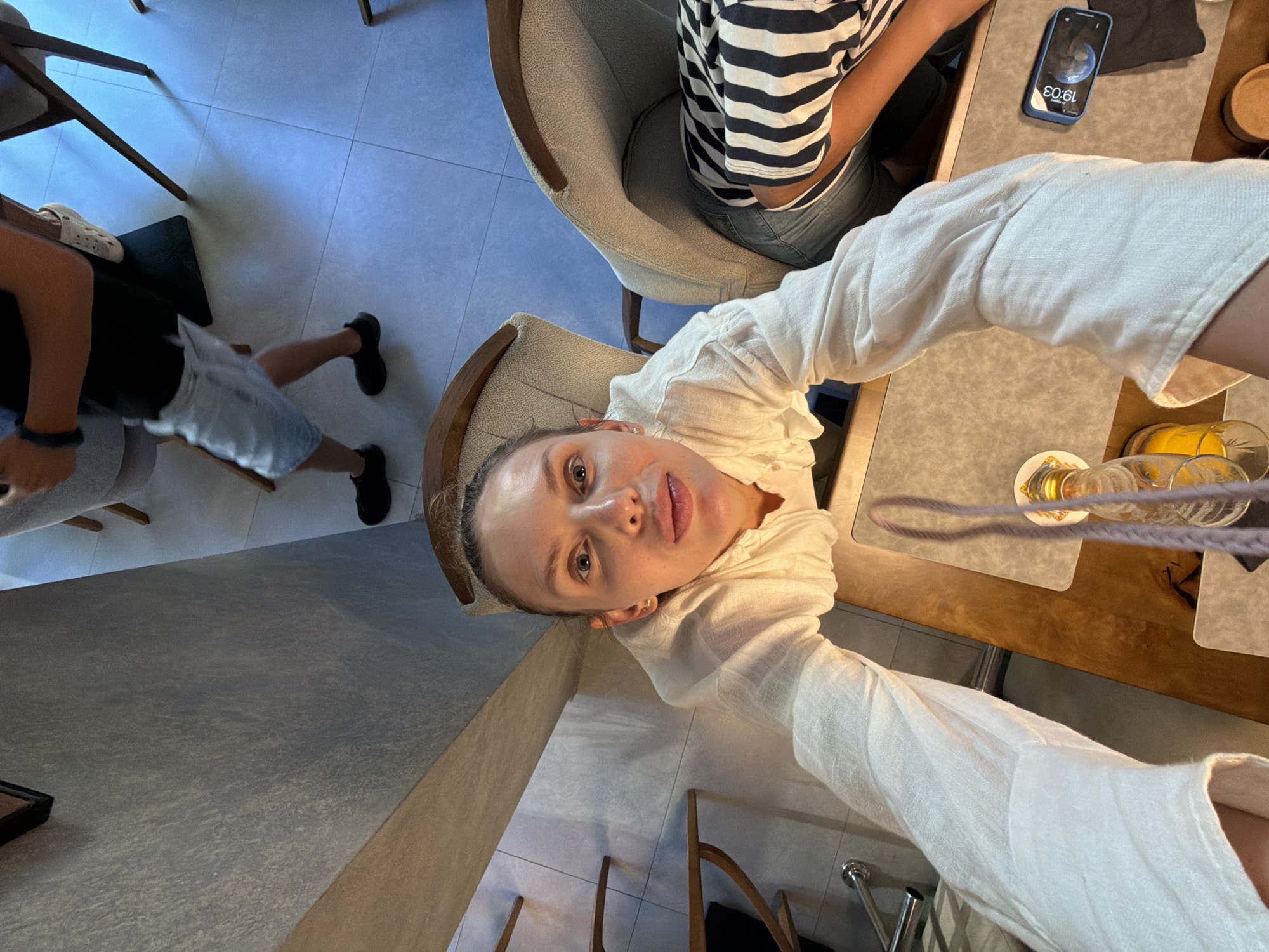 In Budva, Montenegro, a woman is taking a selfie in a well-lit indoor setting, possibly a cafe or restaurant. She is seated, wearing a white linen shirt and blue jeans. Her face is turned towards the camera, and she is looking directly into it with a slightly open mouth. Her hair is pulled back. In the background, another person wearing a black t-shirt and light blue denim shorts is walking away, their back to the camera. The floor is tiled in a light color. To the right, another chair with a light-colored upholstered seat and wooden legs is visible, and a person wearing a black and white striped top and jeans is seated. A wooden table is in the foreground, with a phone displaying the time "19:03" and a glass bottle with a purple string or handle. The lighting suggests it is daytime or early evening.