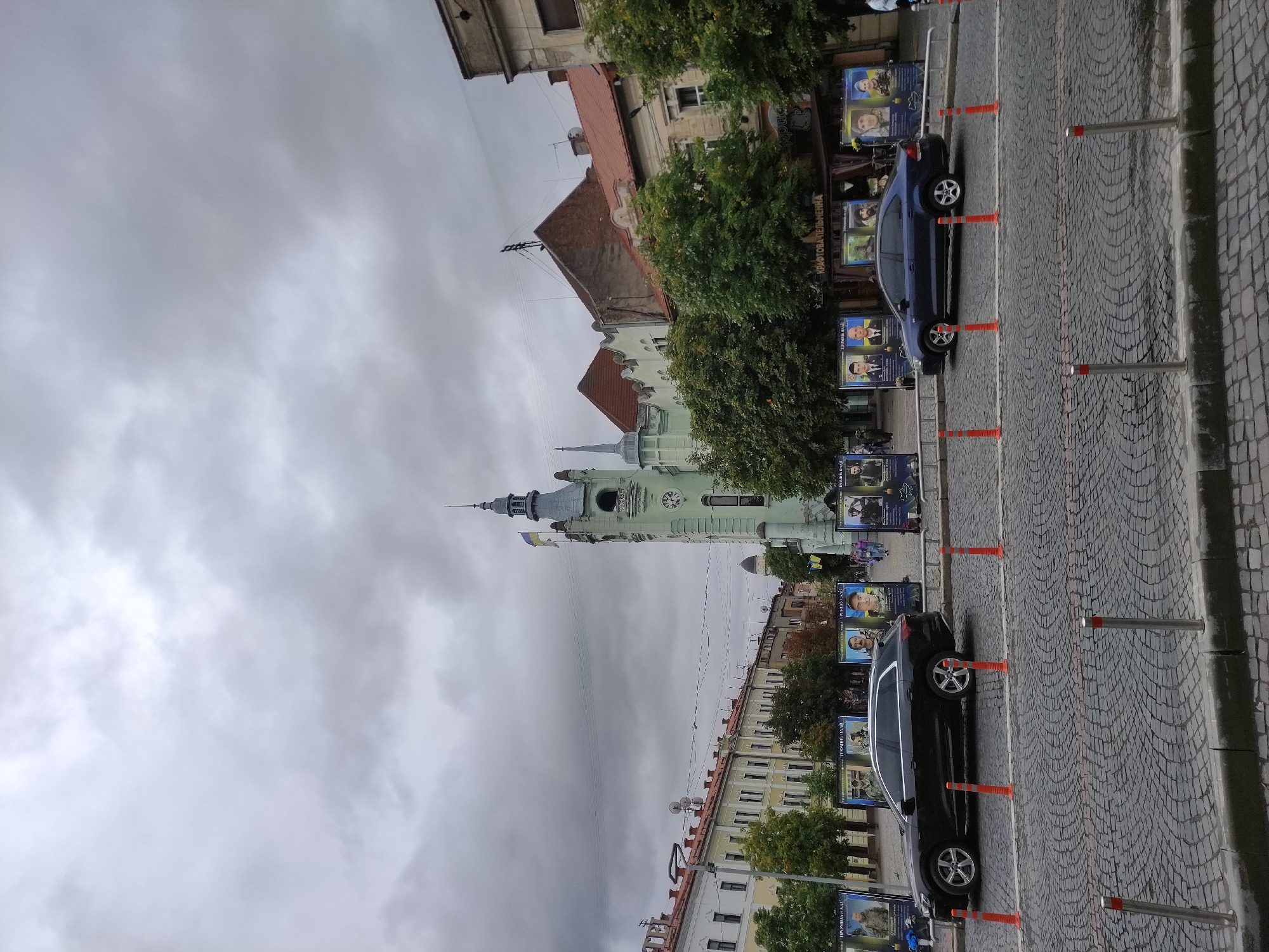 The image depicts a cloudy day in the city of Mukachevo, Ukraine, specifically on the central square, likely near the town hall or a significant historical building. A distinctive light green building with a tower and a clock stands prominently on the right side of the image, adorned with a Ukrainian flag. In front of this building and extending along the street are several large posters, many featuring portraits of individuals and Ukrainian flags. These posters appear to be memorials.
In the foreground, two cars are parked on a cobblestone street separated by orange bollards. A black Audi SUV is parked on the left, and a dark blue sedan is on the right. The street is lined with metal bollards and planters. On the left side of the image, a row of yellow and white buildings with traditional architecture and red-tiled roofs stretches along the street. Trees with green foliage are visible between the buildings and behind the posters. Several people are walking in the background, some near the posters and others in the distance. The overall atmosphere is somber, possibly due to the memorial nature of the displayed posters. The sky is overcast with heavy clouds.