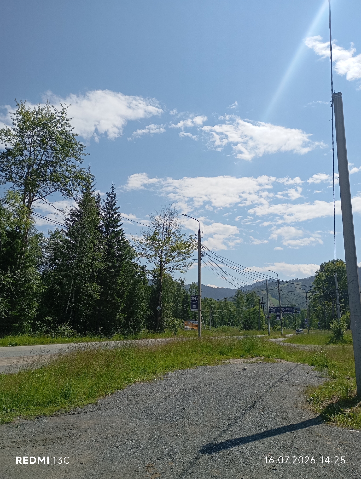 The image captures a sunny day in Baykalsk, Russia, with a clear blue sky dotted with white clouds. In the foreground, a gravelly path leads towards a paved road. Lush green grass and trees line the side of the road, with a dense forest in the background. Mountains are visible in the distance. Several utility poles with electrical wires are present, suggesting infrastructure in the area. A few signs are visible along the road, but their text is not legible. The overall scene depicts a natural, rural setting with some signs of human development.