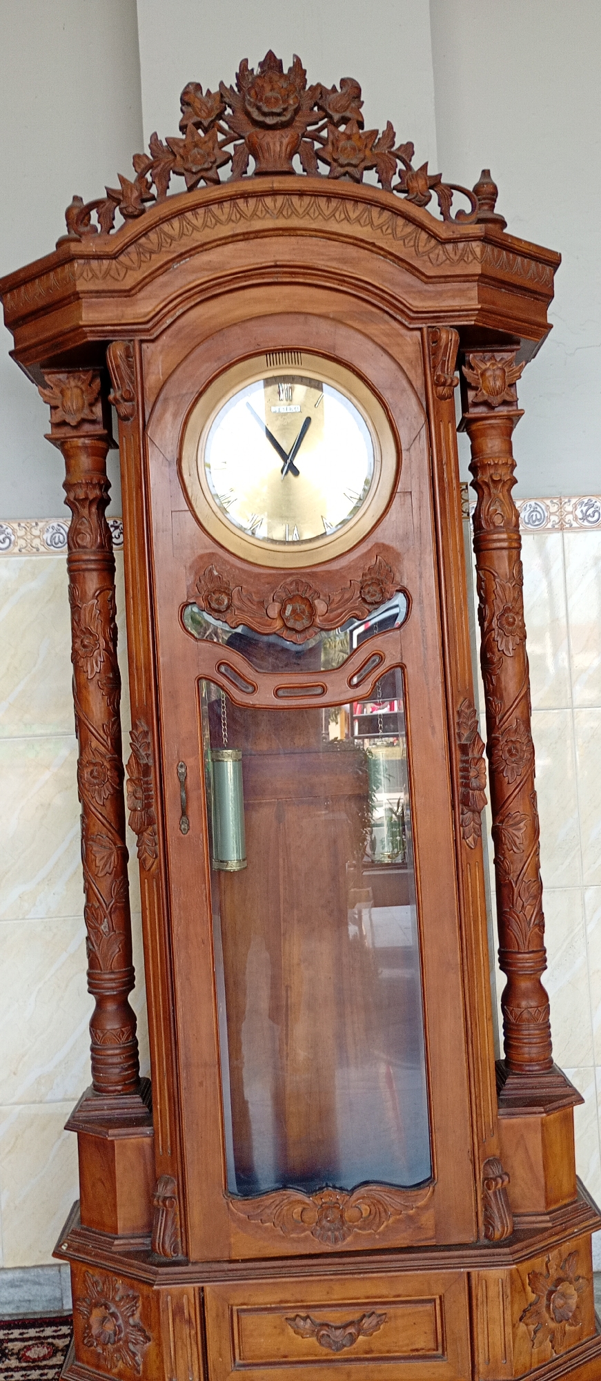 A tall, ornate, wooden grandfather clock stands against a tiled wall. The clock is richly carved with floral motifs and decorative elements. It has a glass-covered face with Roman numerals and visible hands. A pendulum hangs within the glass-fronted body of the clock. The base of the clock is also heavily carved. The background consists of a light-colored wall with a tiled wainscoting. The scene appears to be indoors, possibly in a home or a public space in Probolinggo, Indonesia.