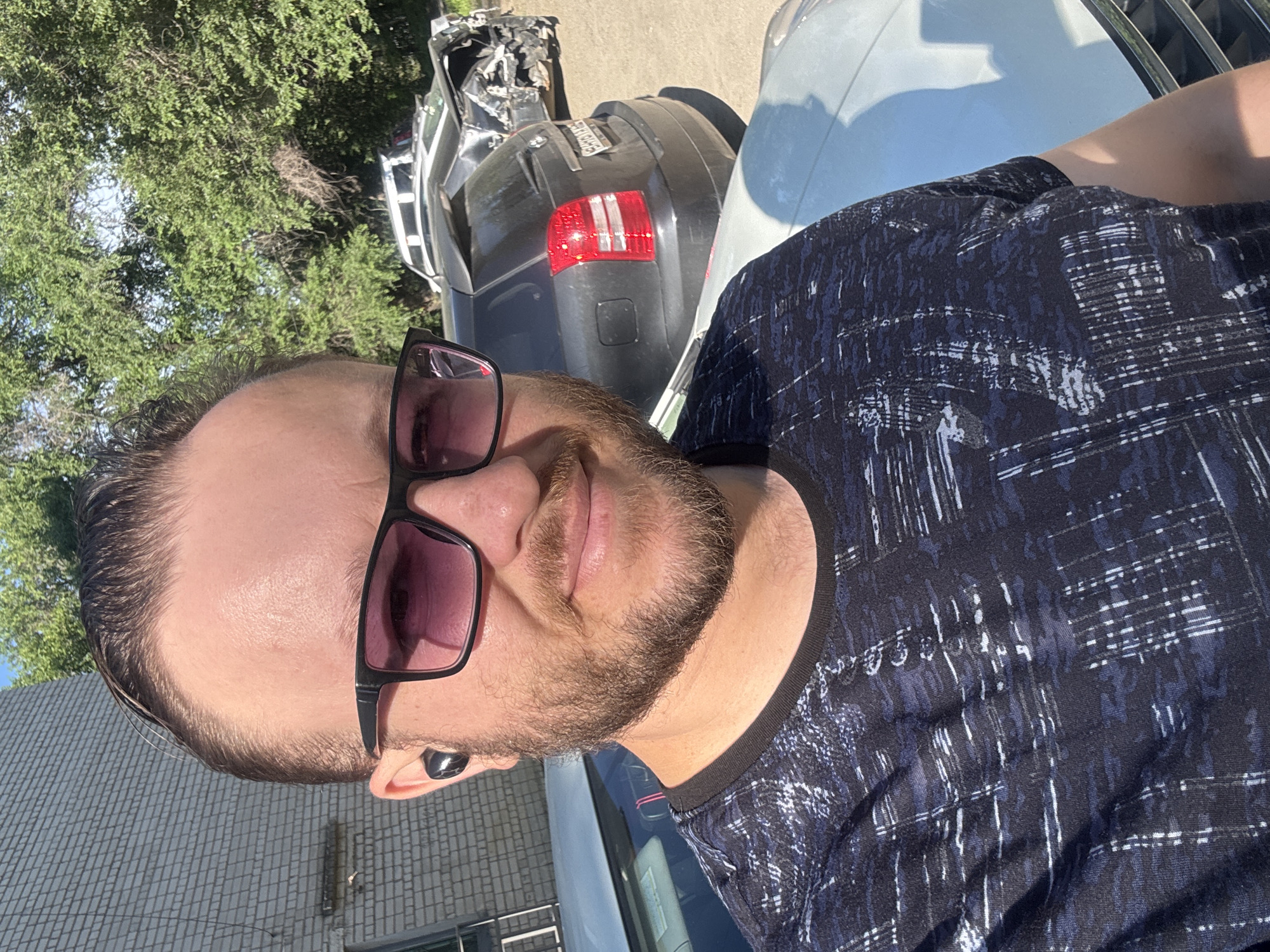 In Blagoveshchensk, Russia, a man with a beard and sunglasses is taking a selfie outdoors. He is wearing a dark t-shirt with a patterned design and wireless earbuds. Behind him, a dark-colored SUV is parked, and further in the background, another car is severely damaged, appearing to have been involved in a collision. The environment suggests a sunny day, with trees and a brick building visible.