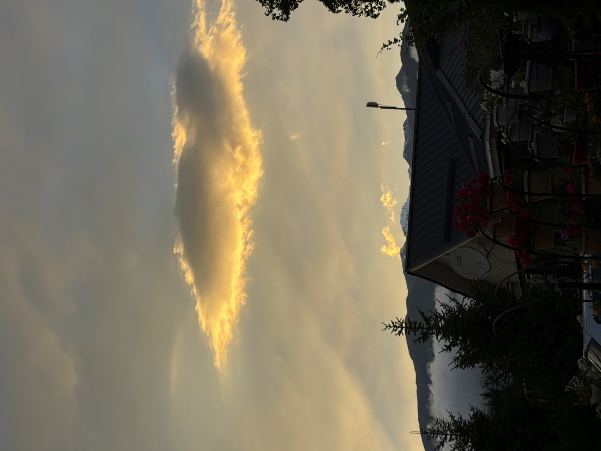 The image shows a twilight scene with a dramatic cloud formation lit by the setting sun. In the foreground, a dark silhouette of a house with a dark roof is visible, along with several evergreen trees and potted plants. Mountains are visible in the background, partially obscured by clouds. A satellite dish is mounted on the side of the house. The overall impression is one of a peaceful, rural setting in Çolpon-Ata, Kyrgyzstan, at dusk.