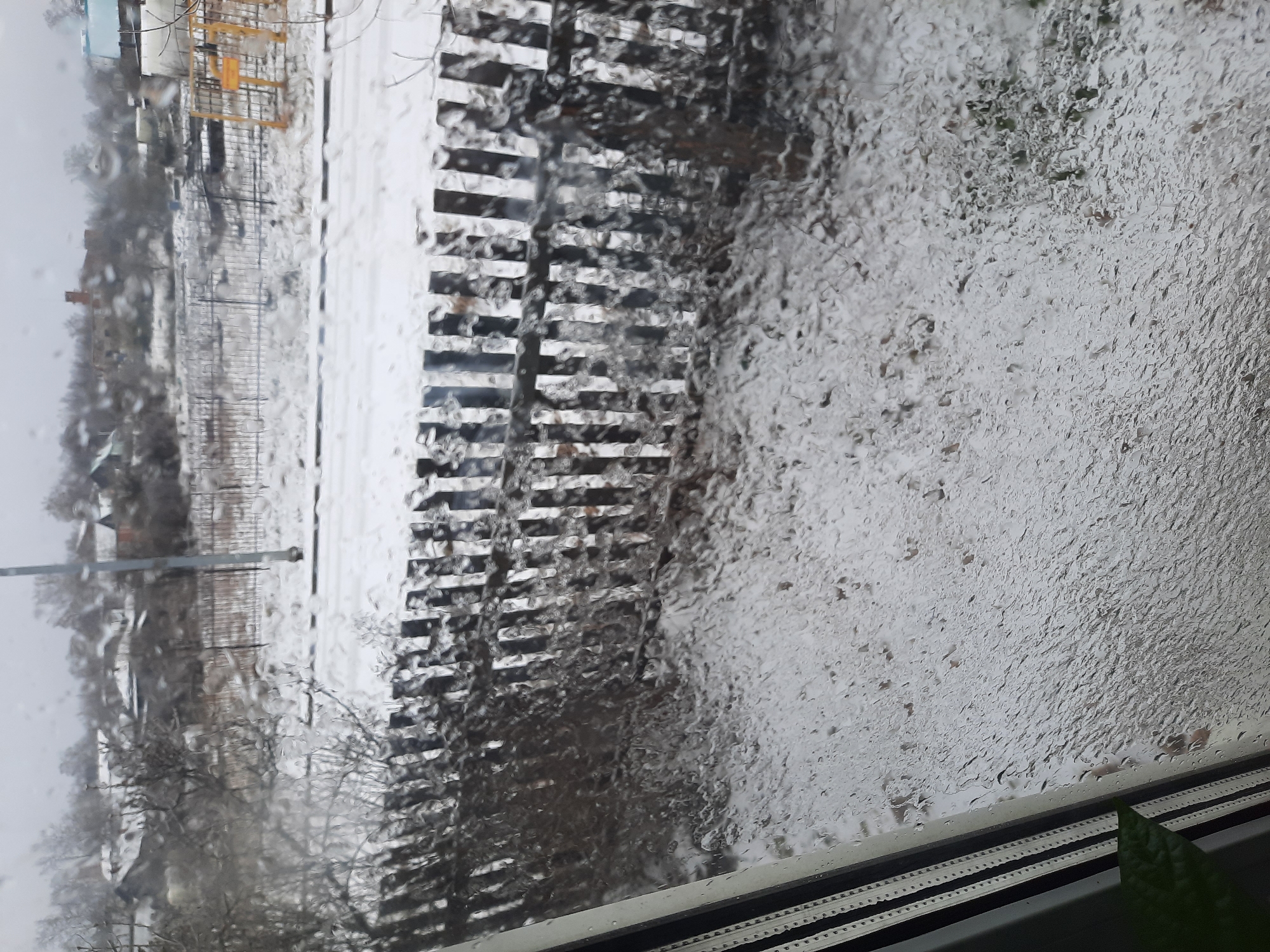 A photograph shows a view through a rain-streaked window of a street scene in Krasnogorskoye, Russia. In the distance, the blurred outlines of buildings and bare trees are visible. A fence runs along the street. The rain on the window obscures details. A yellow playground structure is visible.