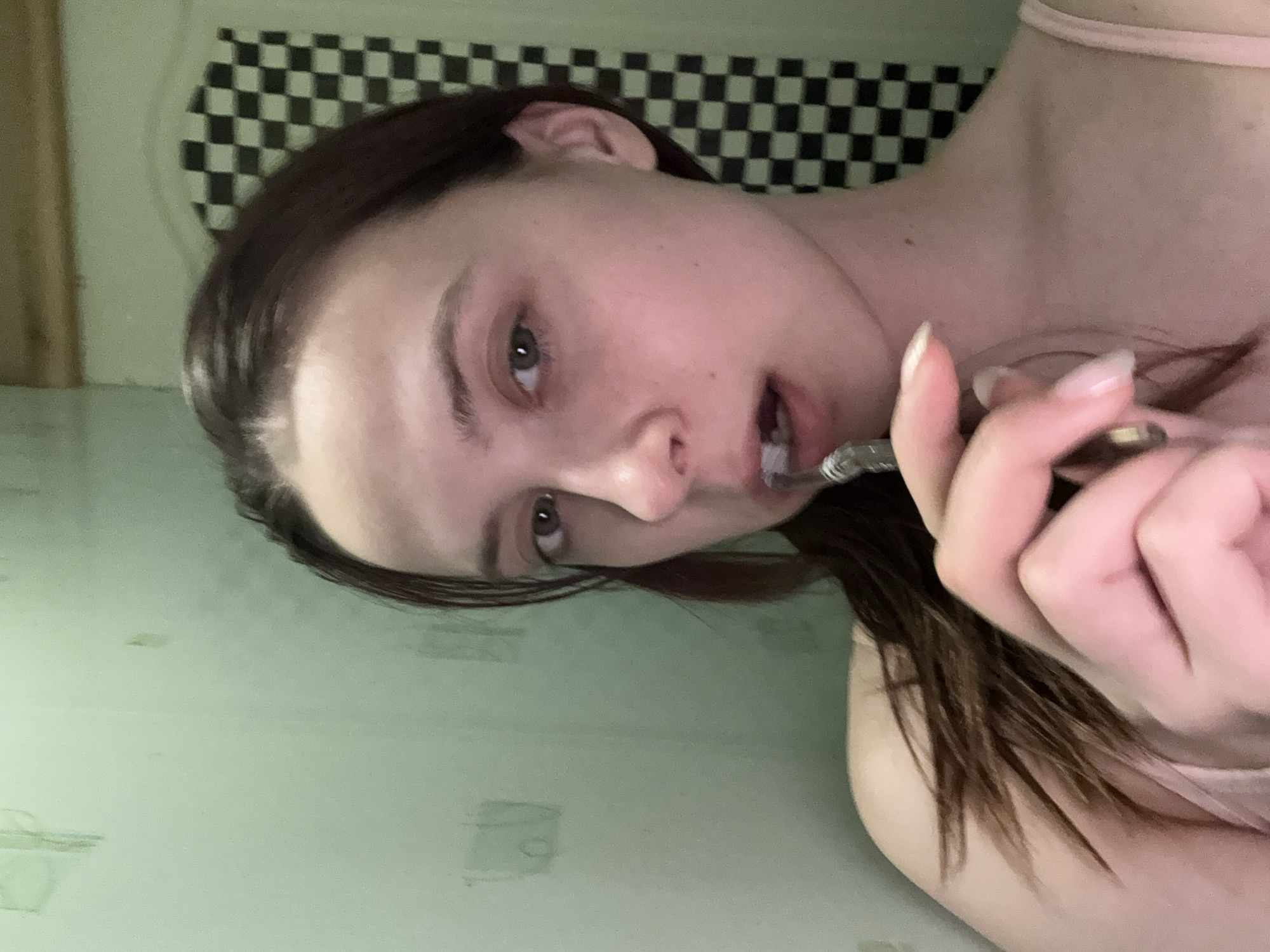 A young, light-skinned woman is seen in a close-up, rotated view. She has dark hair and fair skin. She is holding a small silver object in her hand, and it appears to be near her mouth. The background shows a light green wall with some smudges or stains. Above, there is a black and white checkered pattern visible on the wall. The location is not discernible as being in the city of Прикамский, Russia.