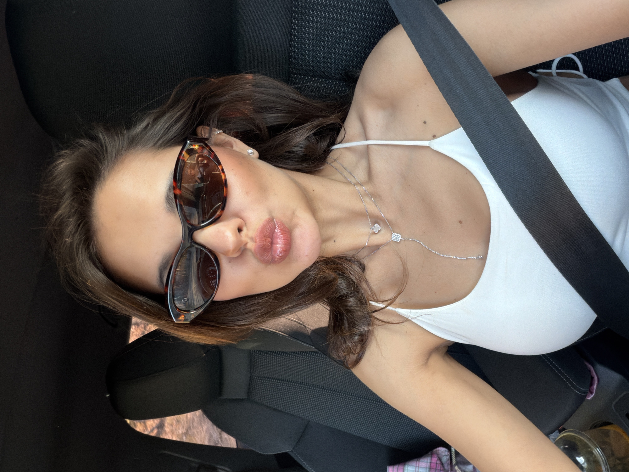 A woman with brown hair and sunglasses is sitting in a car, wearing a white tank top and a silver necklace. She has a seatbelt on and is pouting her lips. The car seat is black. There are no indications of the location being Krasnoyarsk, Russia.